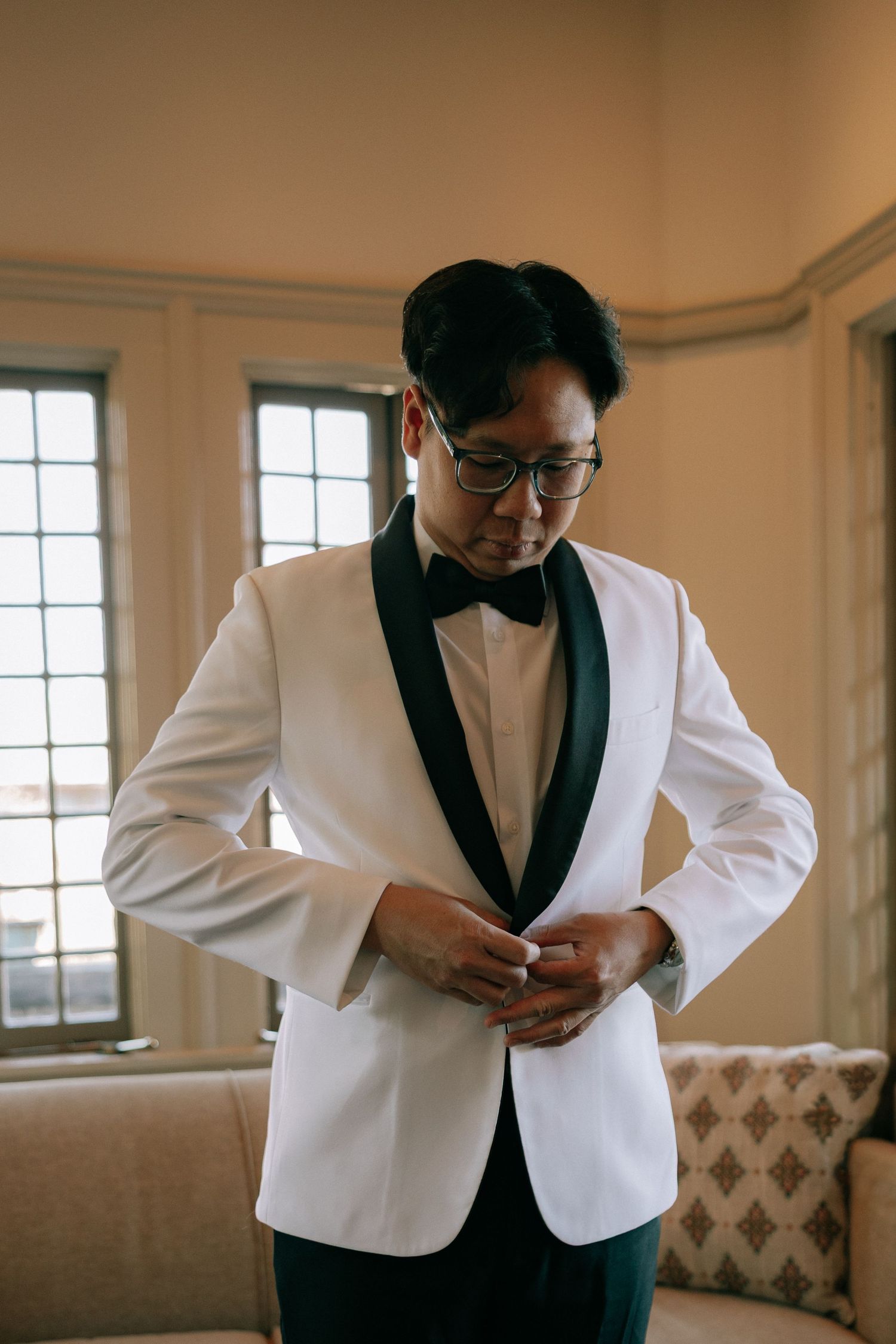 Groom in white tuxedo jacket with black lapels and bow tie buttoning his jacket before wedding ceremony.