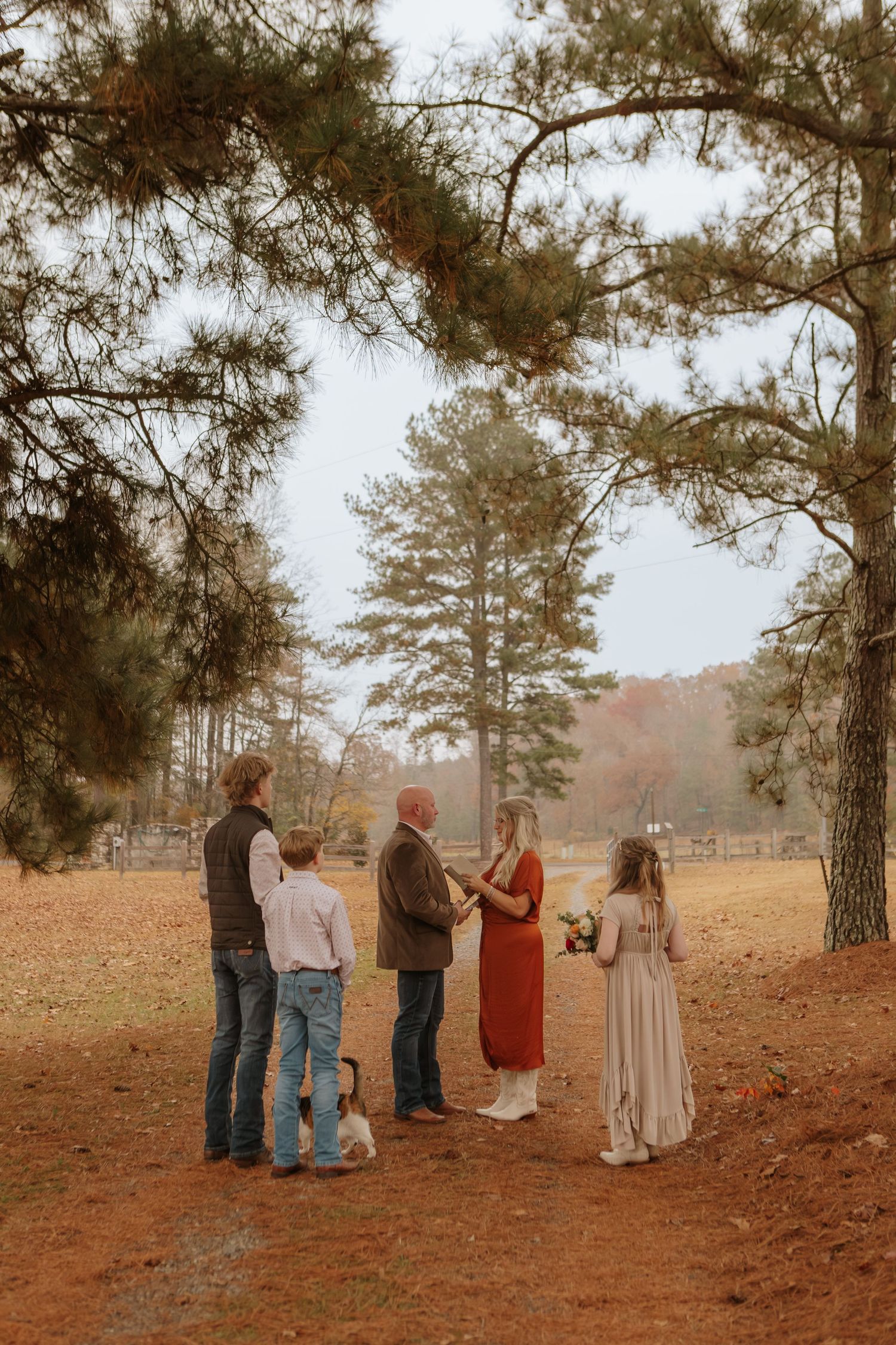 Small intimate outdoor wedding ceremony under pine trees with family gathering on red clay ground in autumn.