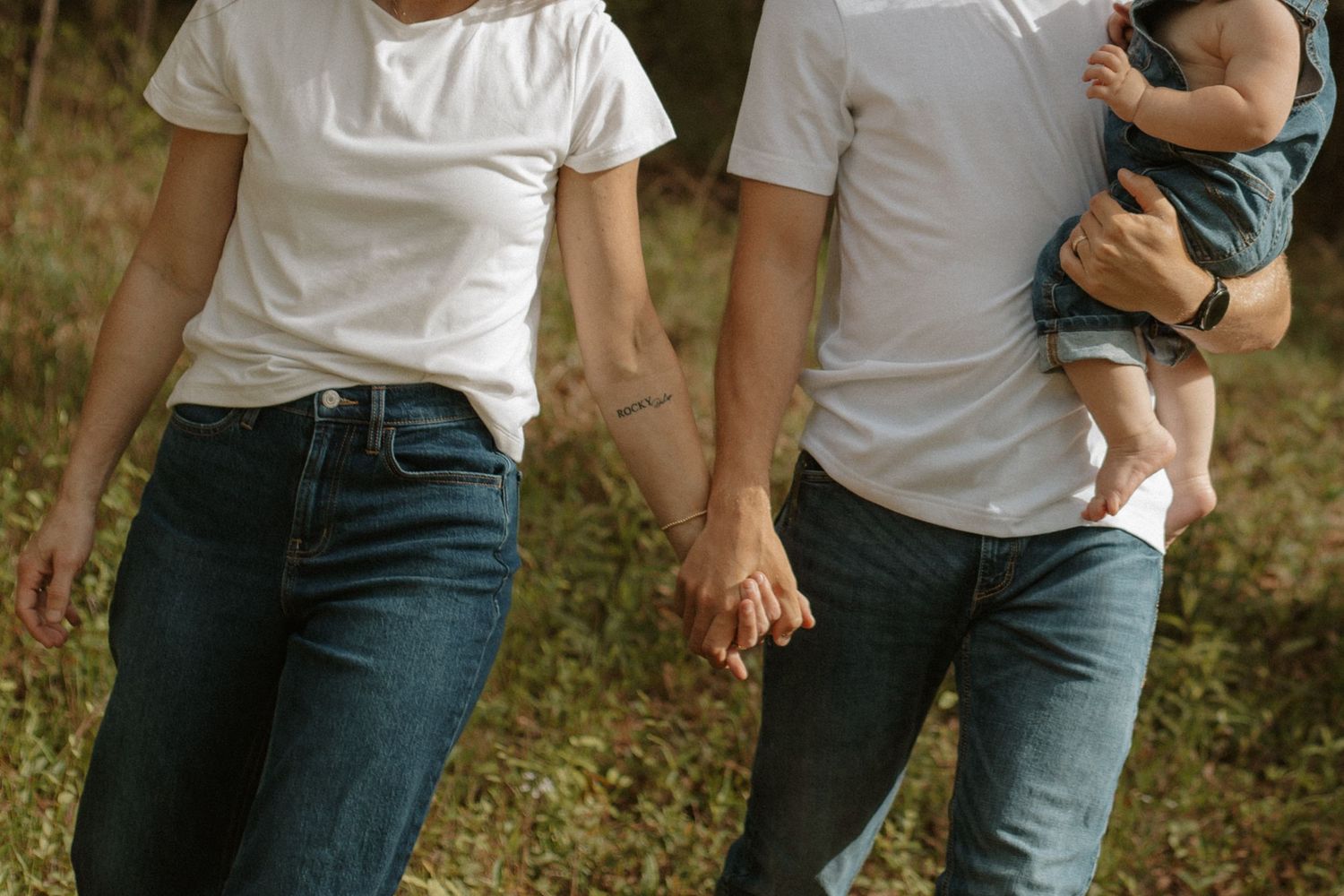 Couple in white tees and jeans hold hands while man carries baby through green forest path.