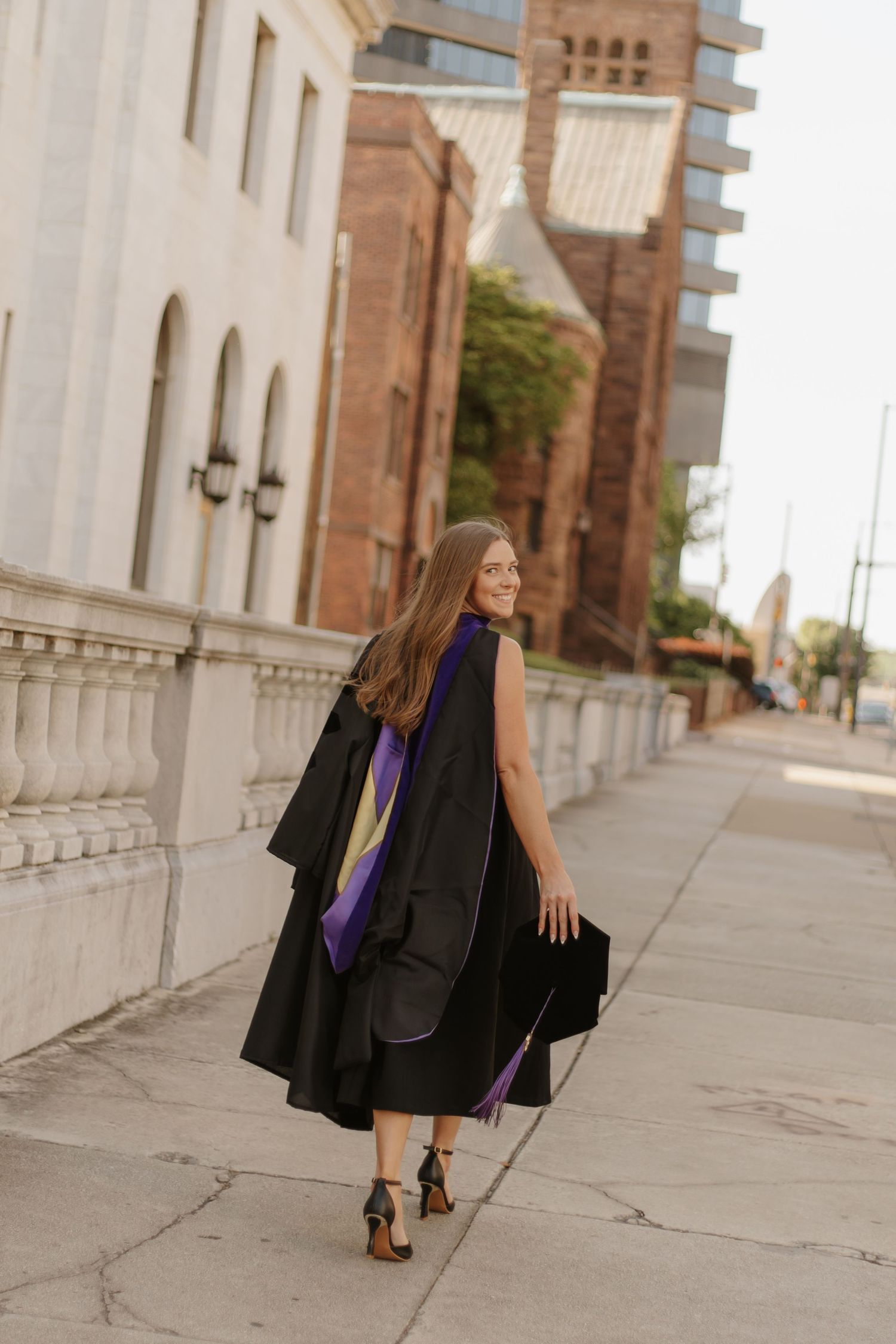 Graduate in black gown with purple stole walking along urban sidewalk near historic building.