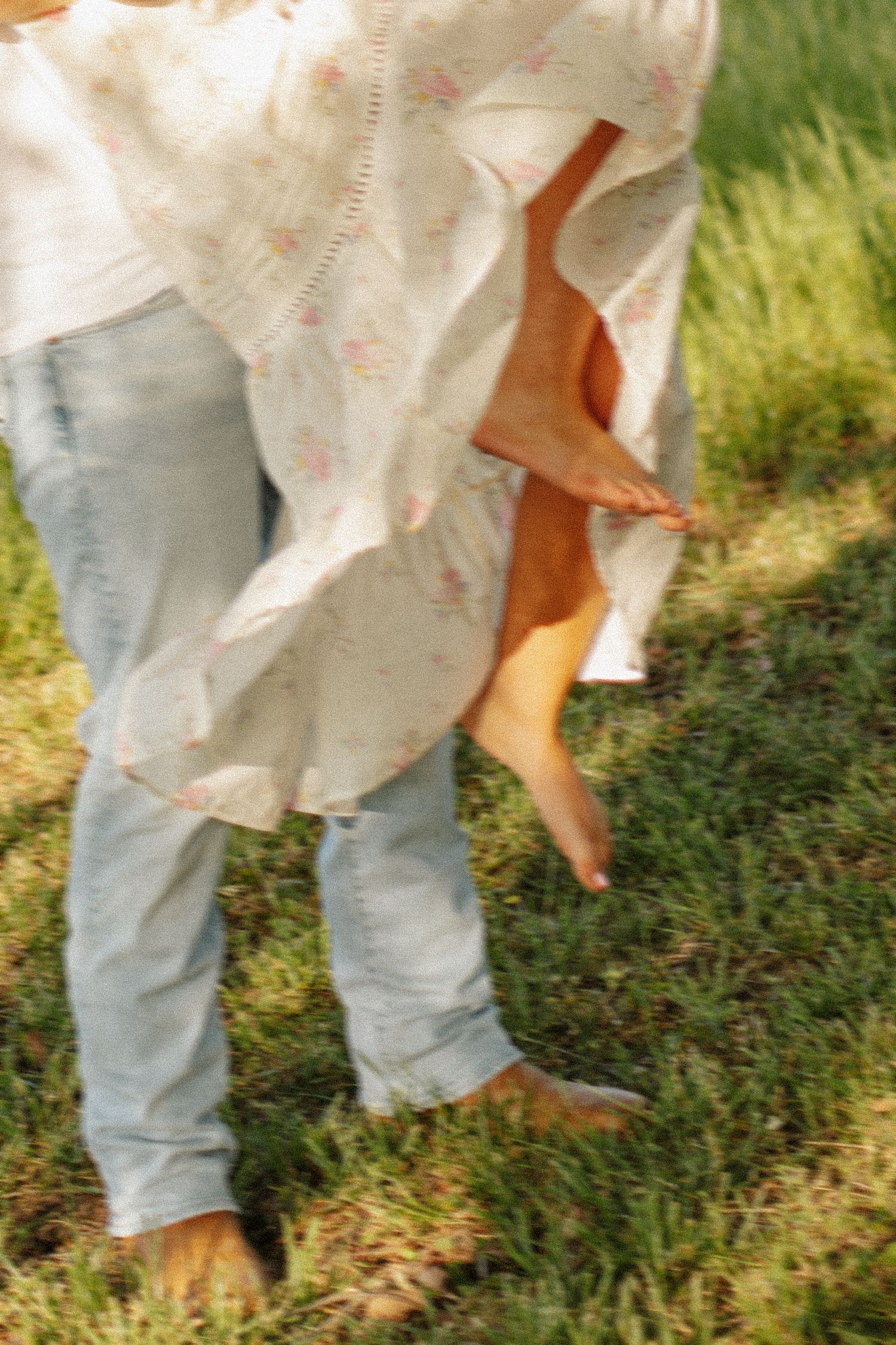Adult carrying barefoot adult outdoors on lush green grass, wearing floral dress and jeans.