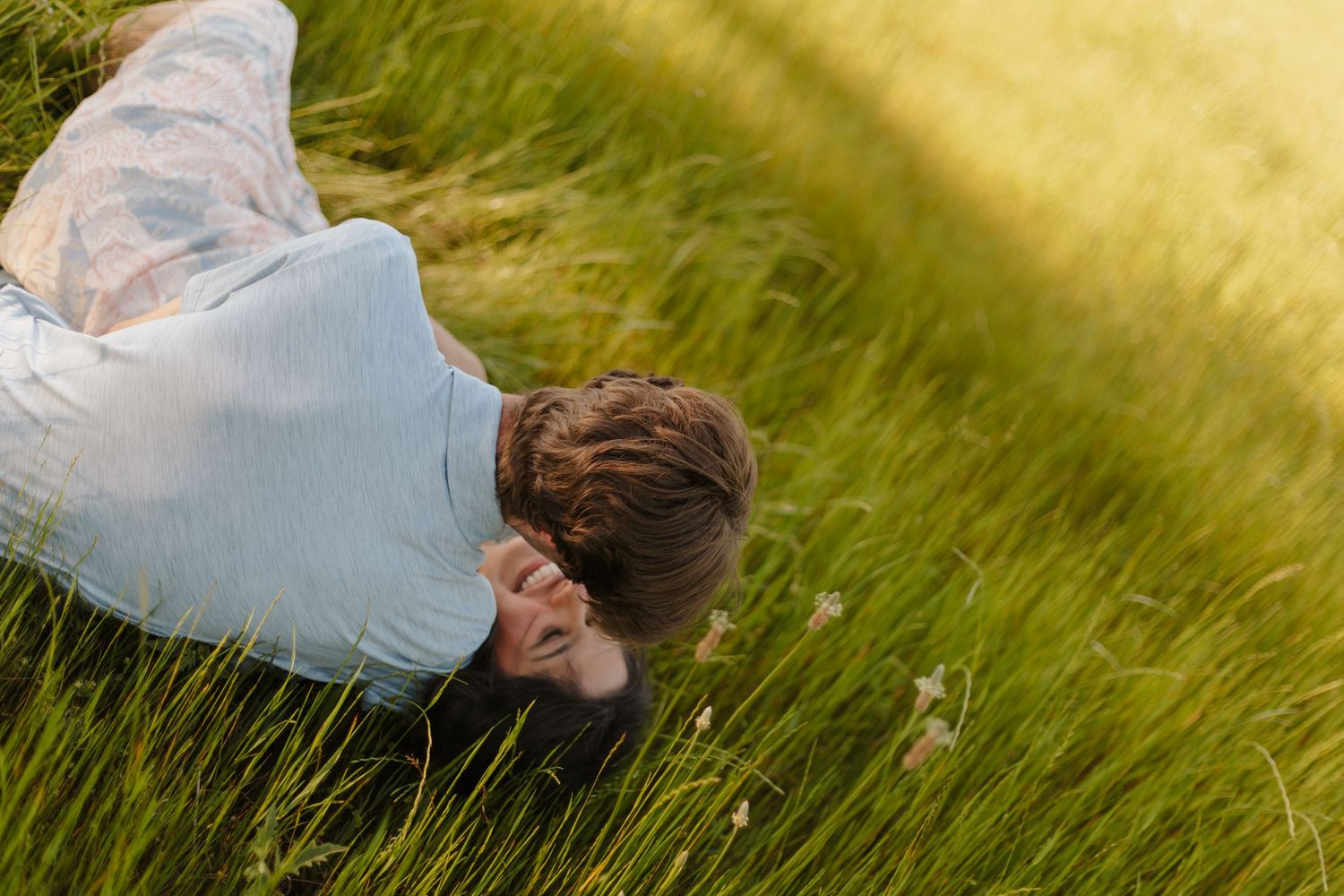 Couple lying together and laughing in tall green grass during a warm sunny day outdoors.