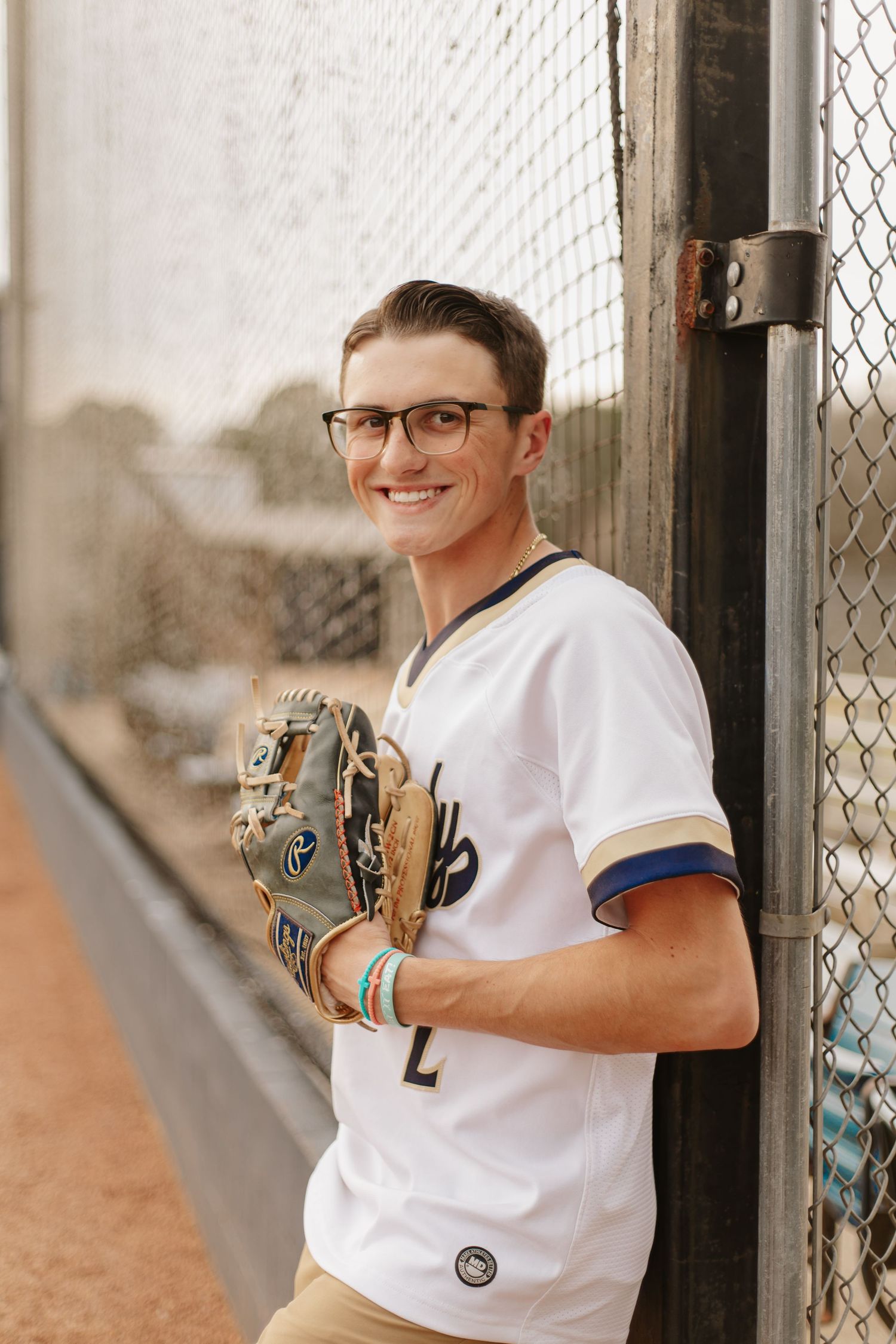 Smiling teen boy in baseball jersey and glasses holds glove while leaning against chain-link fence.