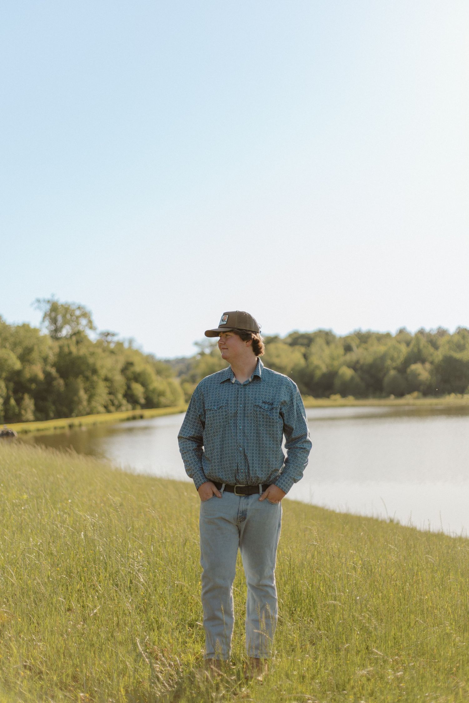 Young man in plaid shirt and cap stands in green field beside a calm lake on a sunny day.
