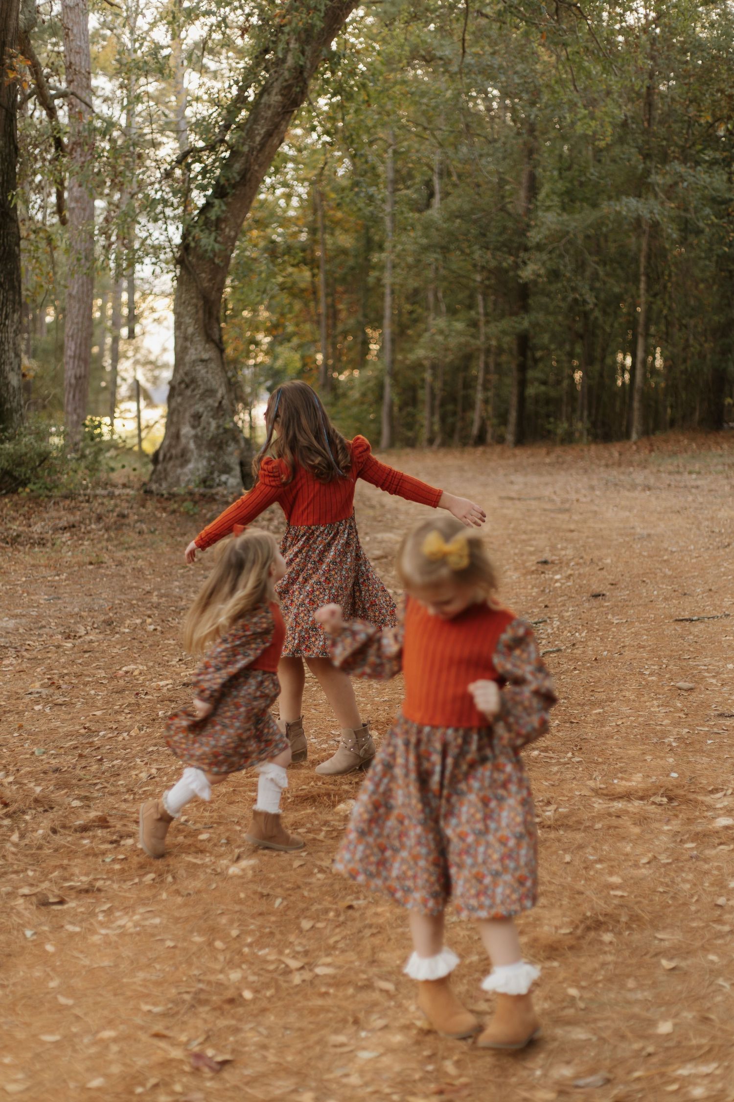 Three girls in floral dresses and rust sweaters spin on a leaf-covered forest path.