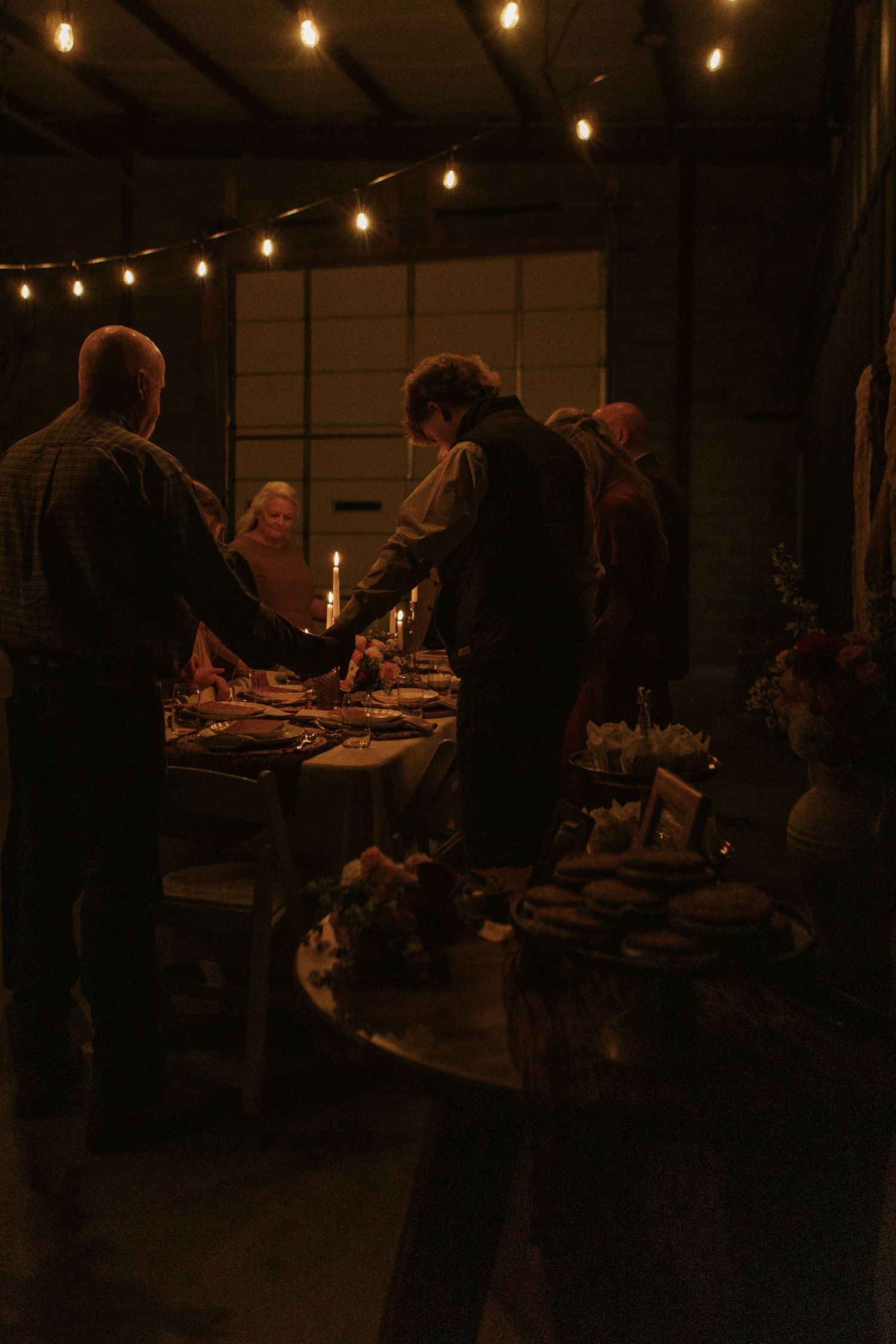 Guests holding hands around a candlelit dinner table with string lights in a rustic dark venue.