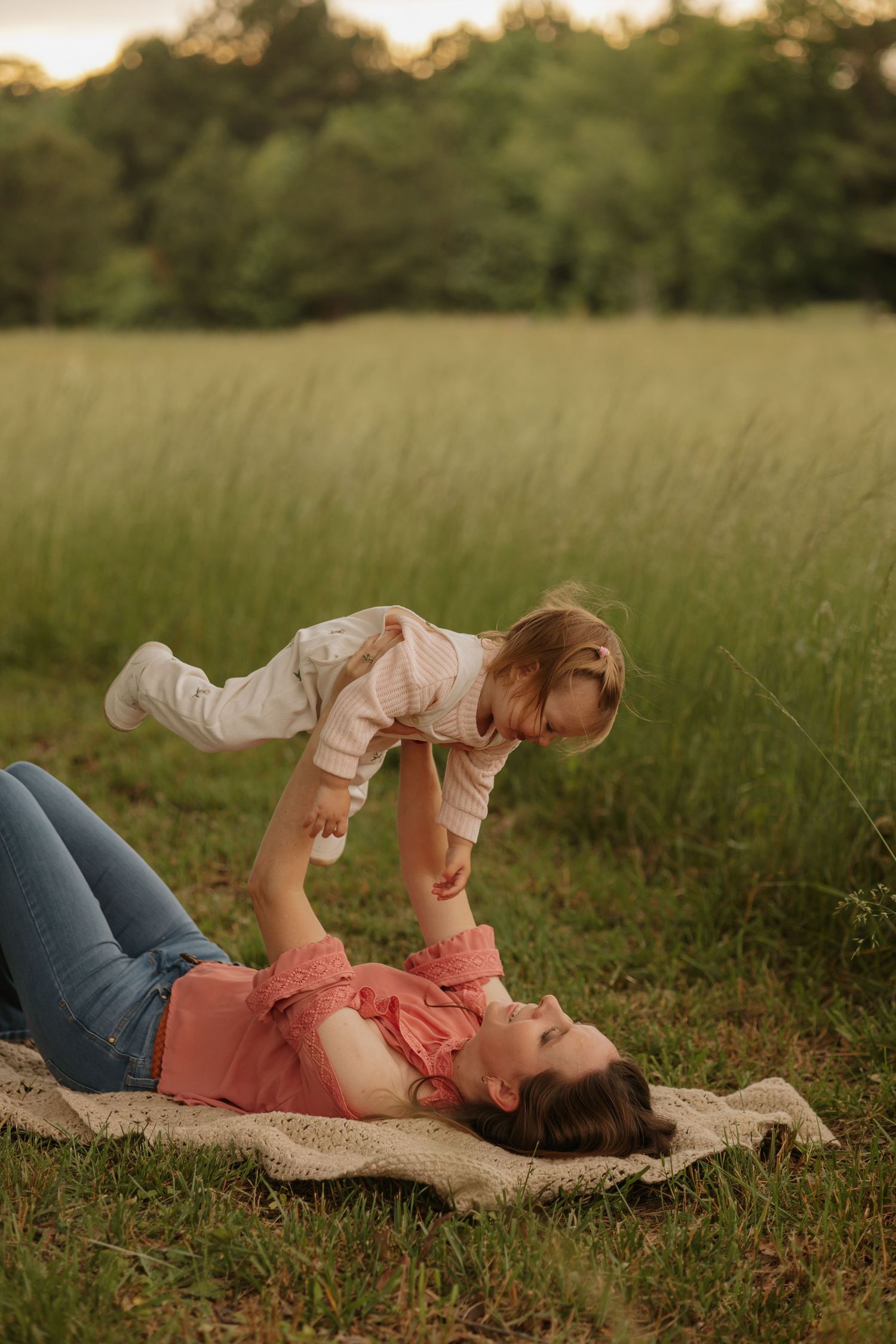 A mother lies on a blanket in a green field playfully lifting her laughing toddler into the air.
