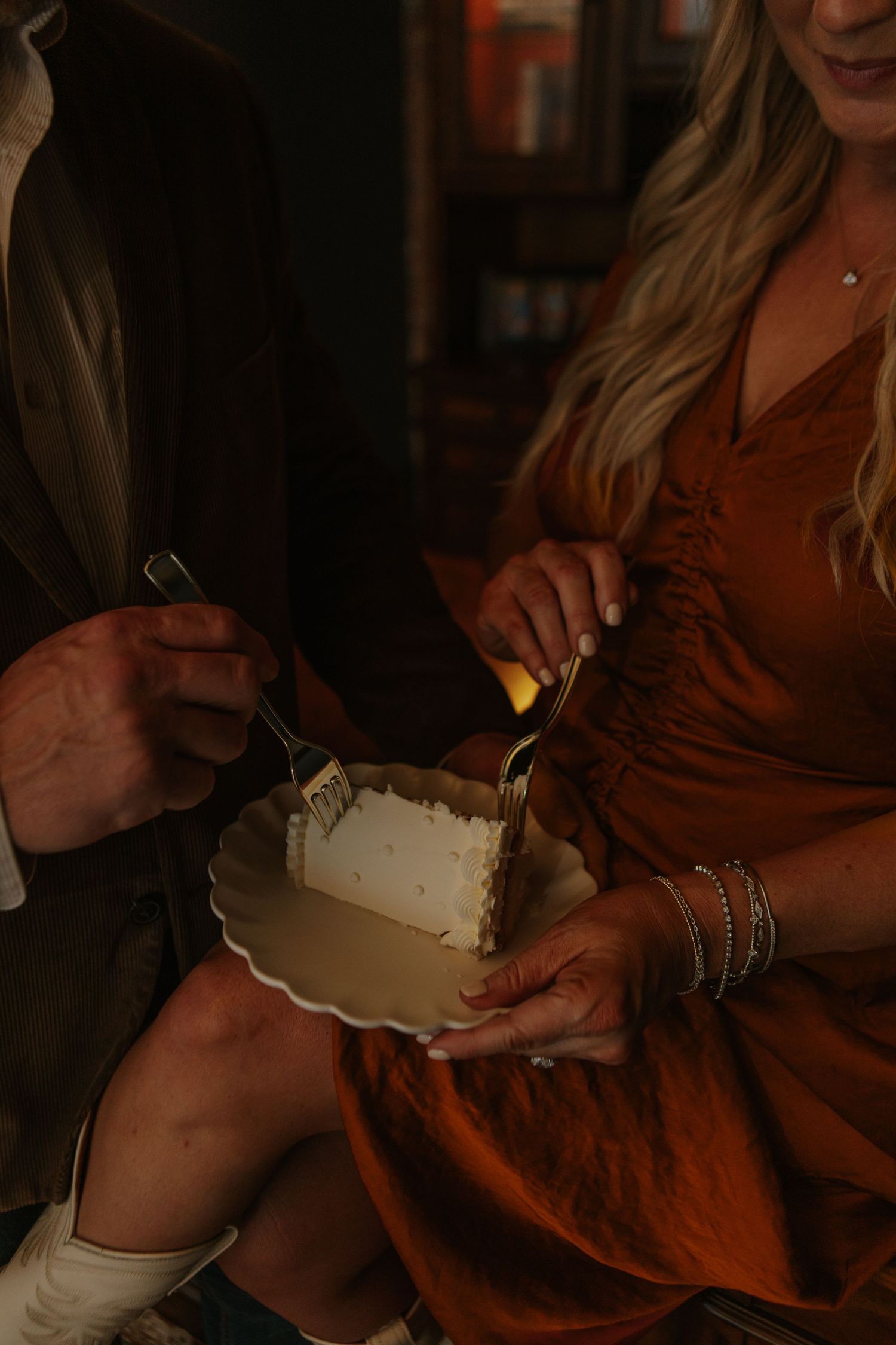 Couple sharing a slice of white wedding cake, bride wearing rust orange dress with gold bracelets.