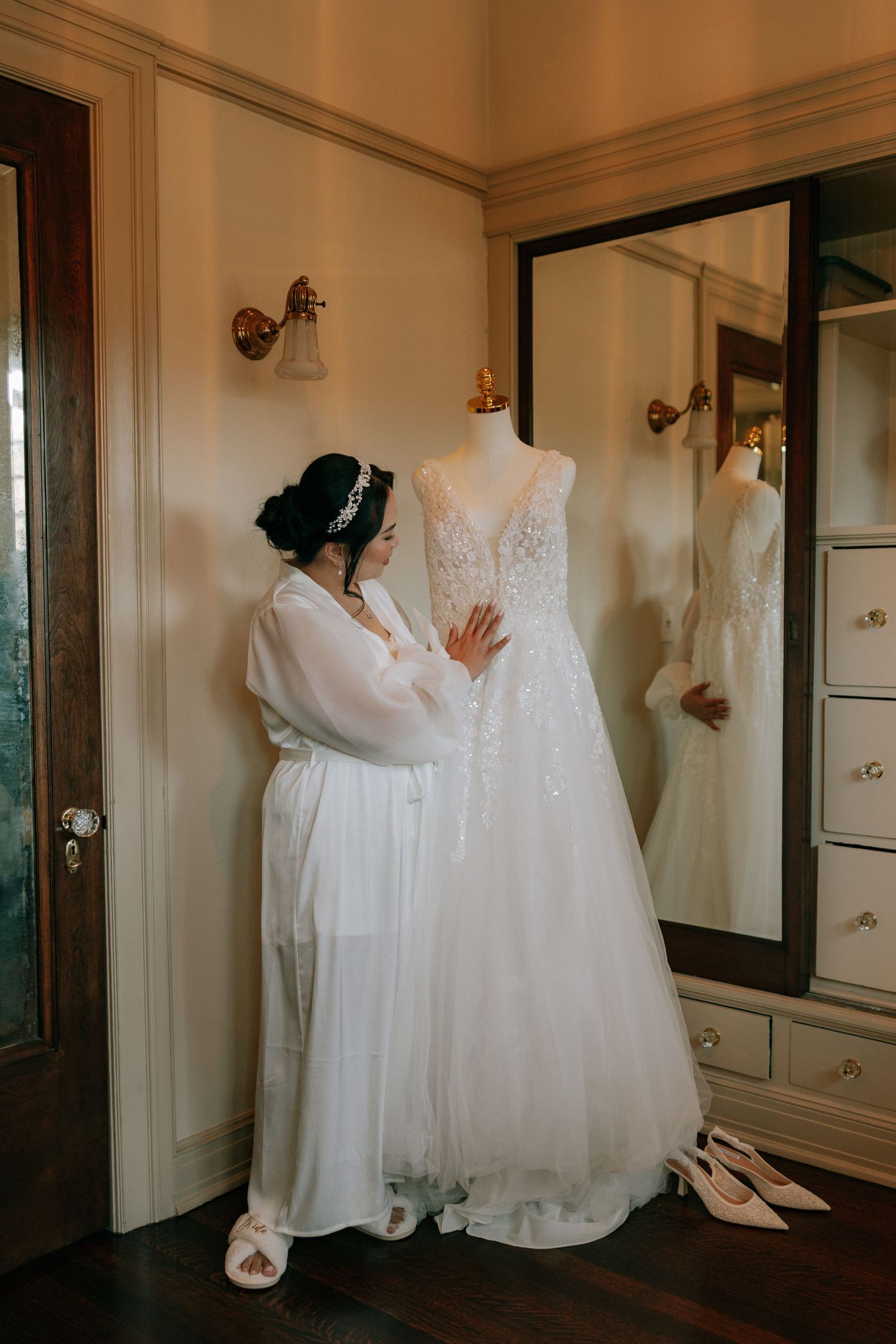 Bride in white robe admires wedding gown on mannequin in elegant dressing room with mirror.