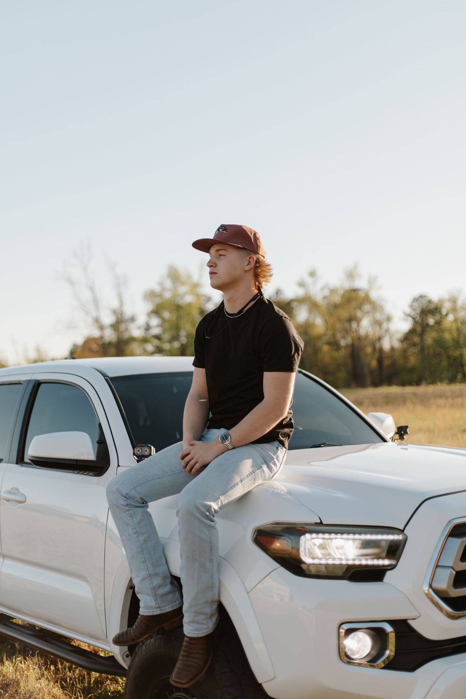 Young man in black shirt and cap sitting on hood of white Toyota Tacoma at sunset.