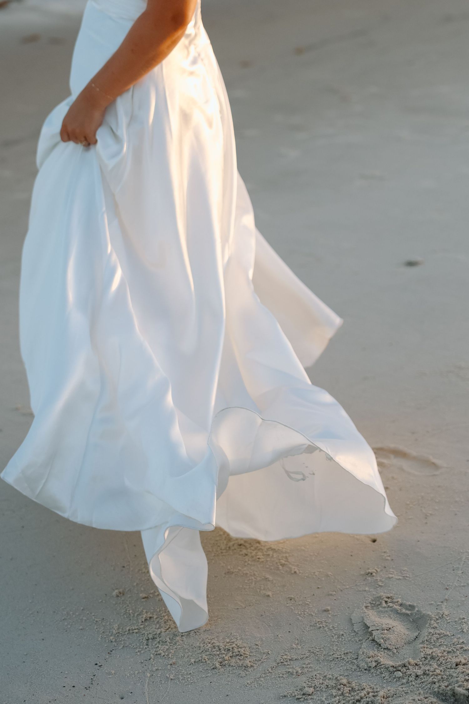 Bride in flowing white wedding gown walks on sandy beach with dress billowing in the breeze.