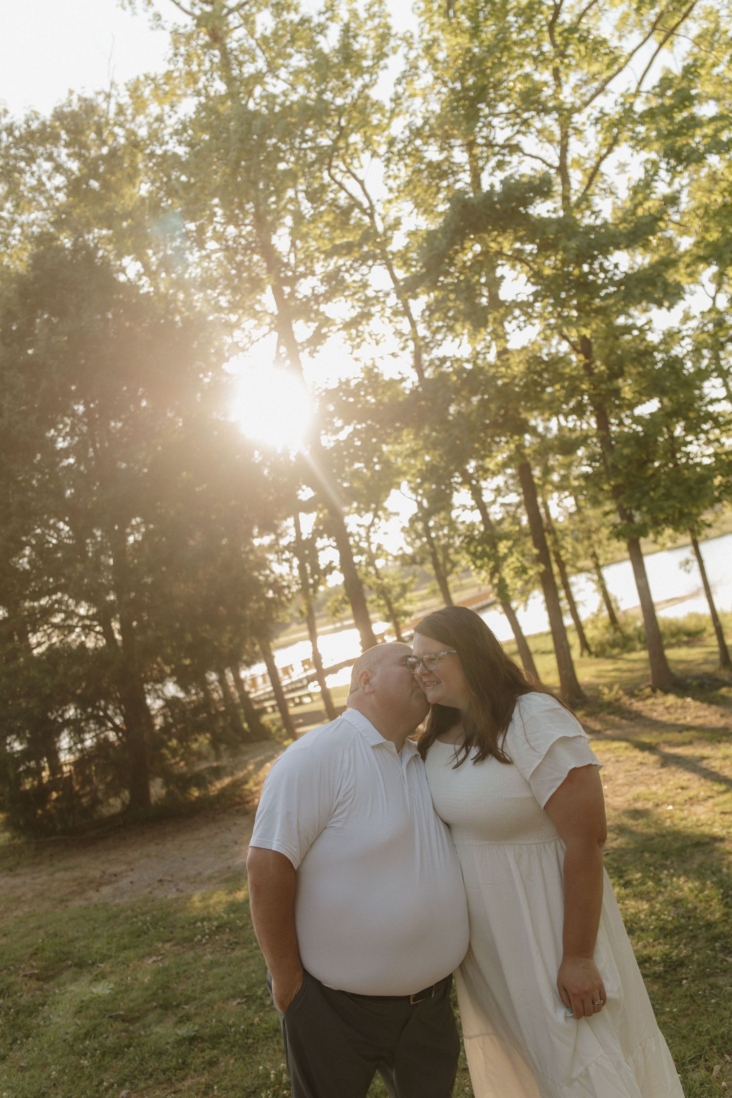Couple sharing a tender kiss outdoors in golden sunset light surrounded by tall green trees.