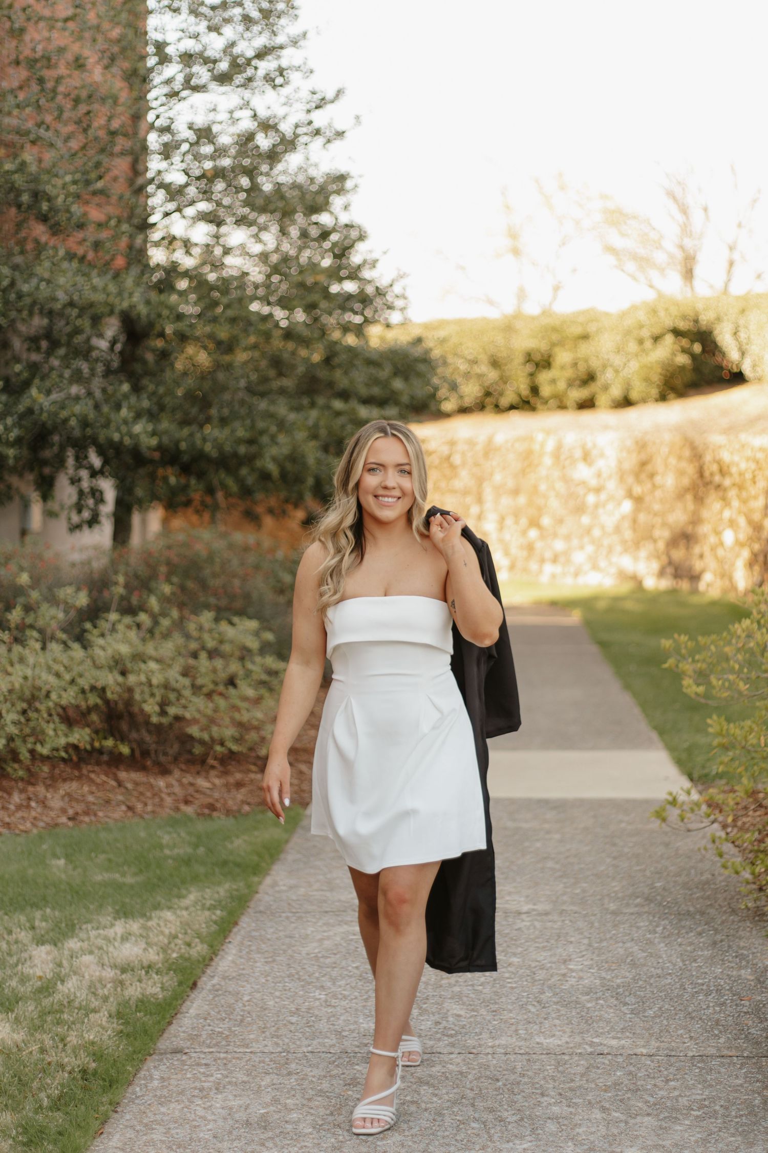 Smiling blonde woman in white strapless dress carries black blazer while walking on sunny pathway.