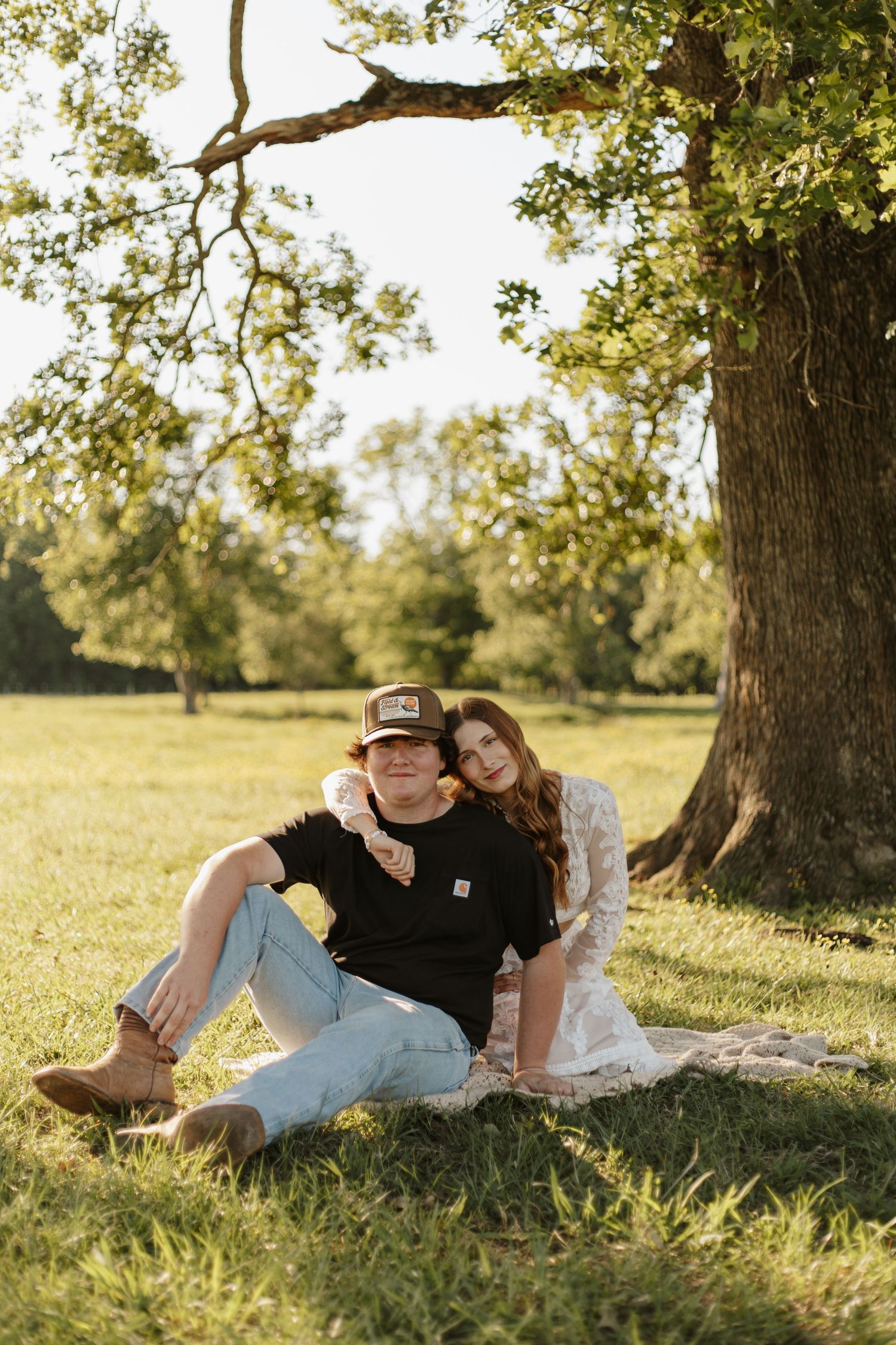 Couple sitting together under a large tree in a sunny green field, woman leaning on mans shoulder.