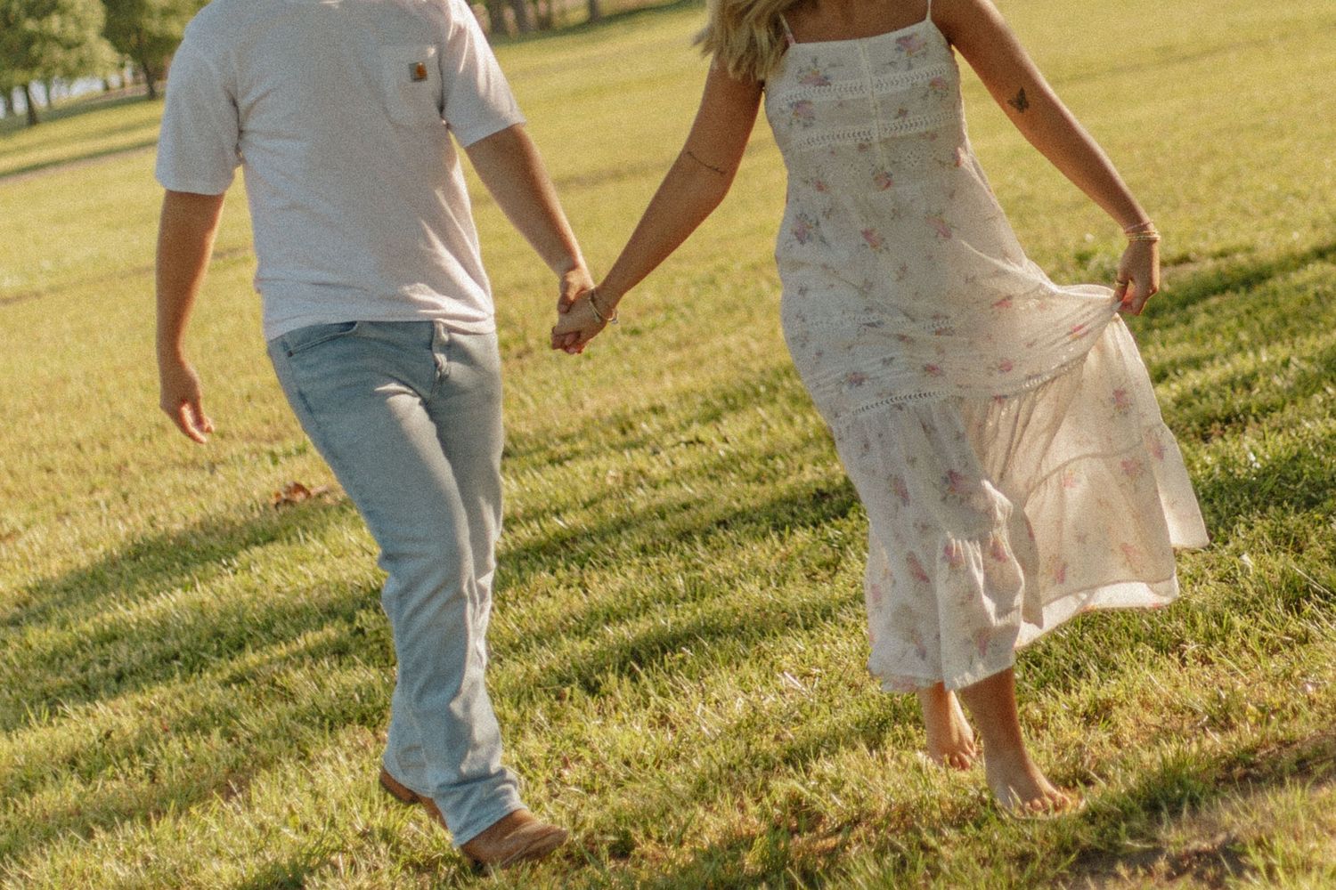 Couple holding hands and walking barefoot through a sunny green grass field together.