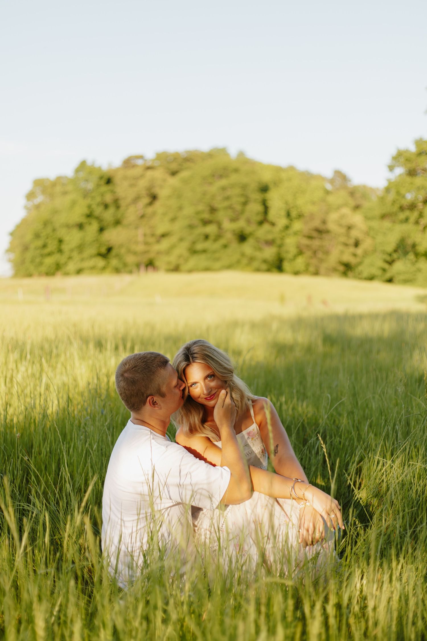 Romantic couple sitting together in a sunlit green field, sharing an intimate moment with trees in the background.