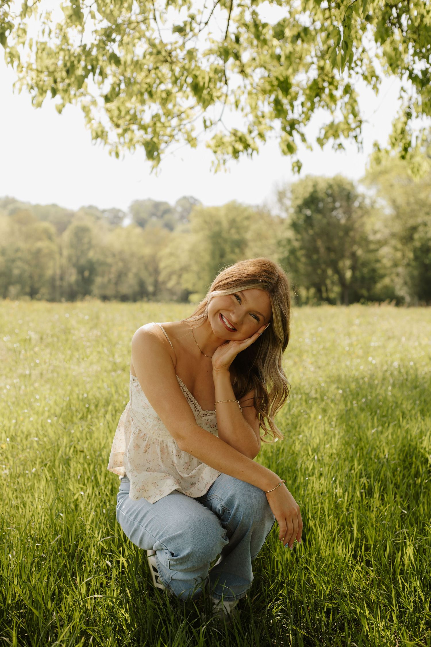 Smiling woman in lace top and jeans crouching in a lush green meadow.