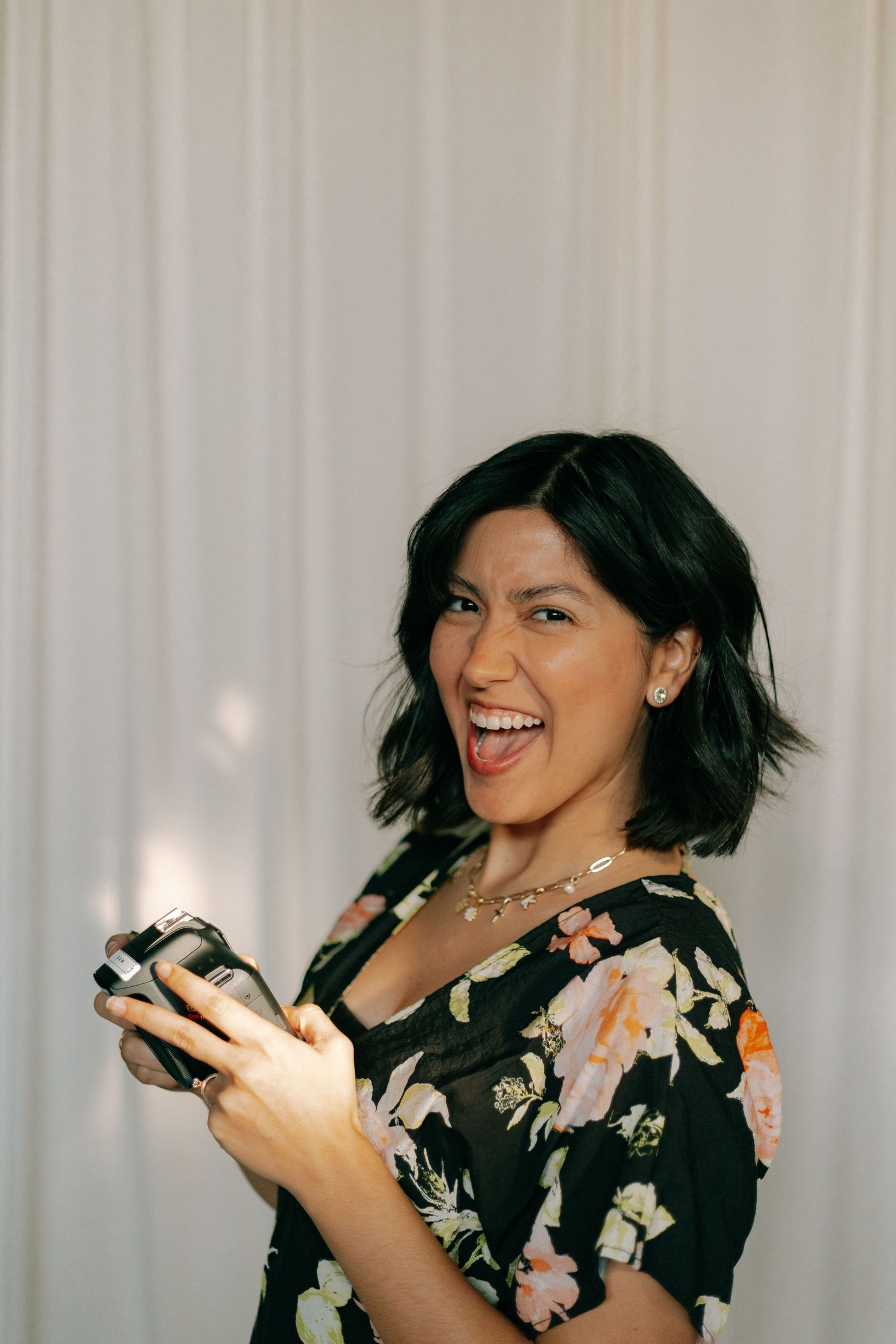 Smiling woman with short dark hair holding a Canon camera wearing a floral dress.