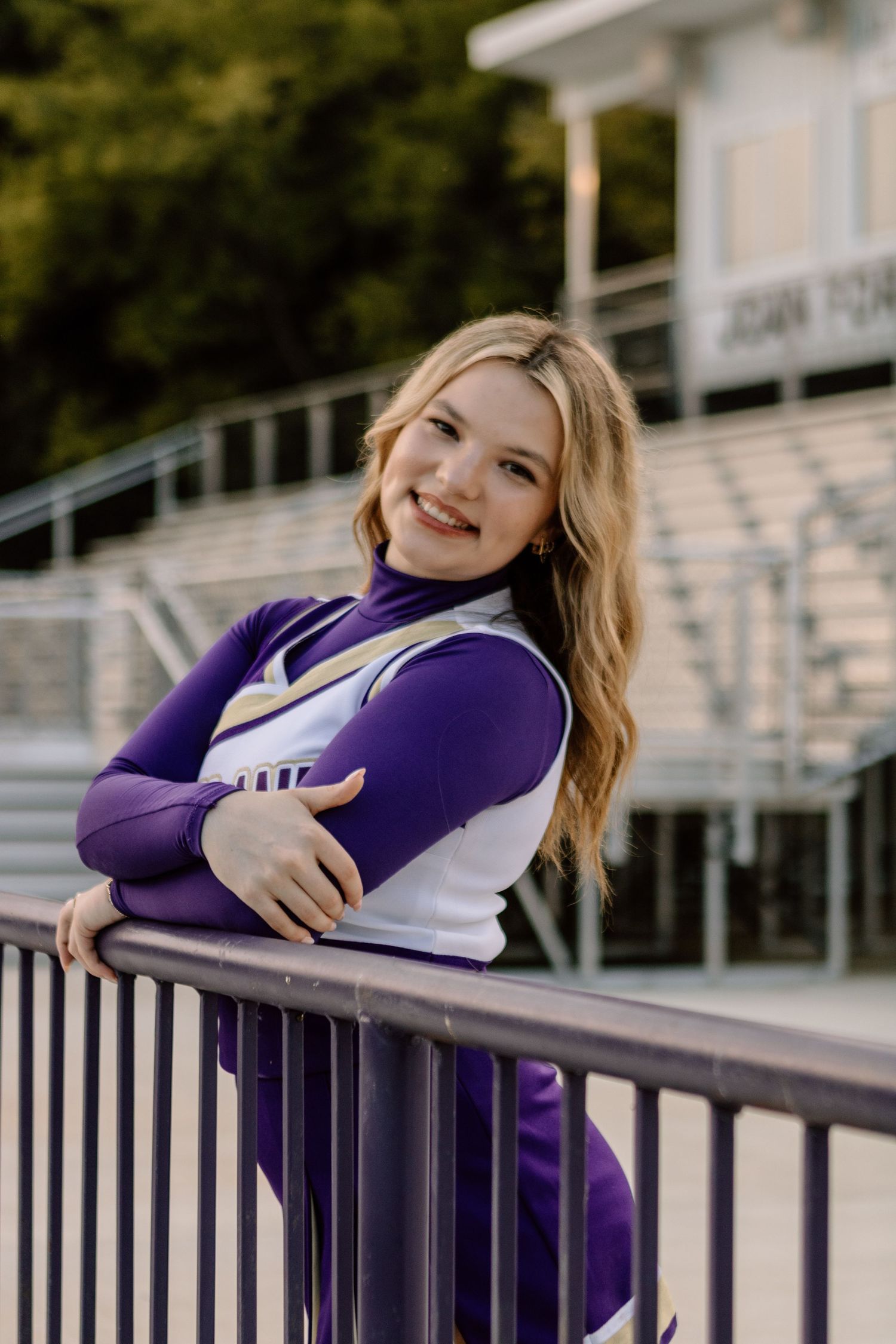 Blonde cheerleader in purple uniform smiling while leaning on stadium railing.