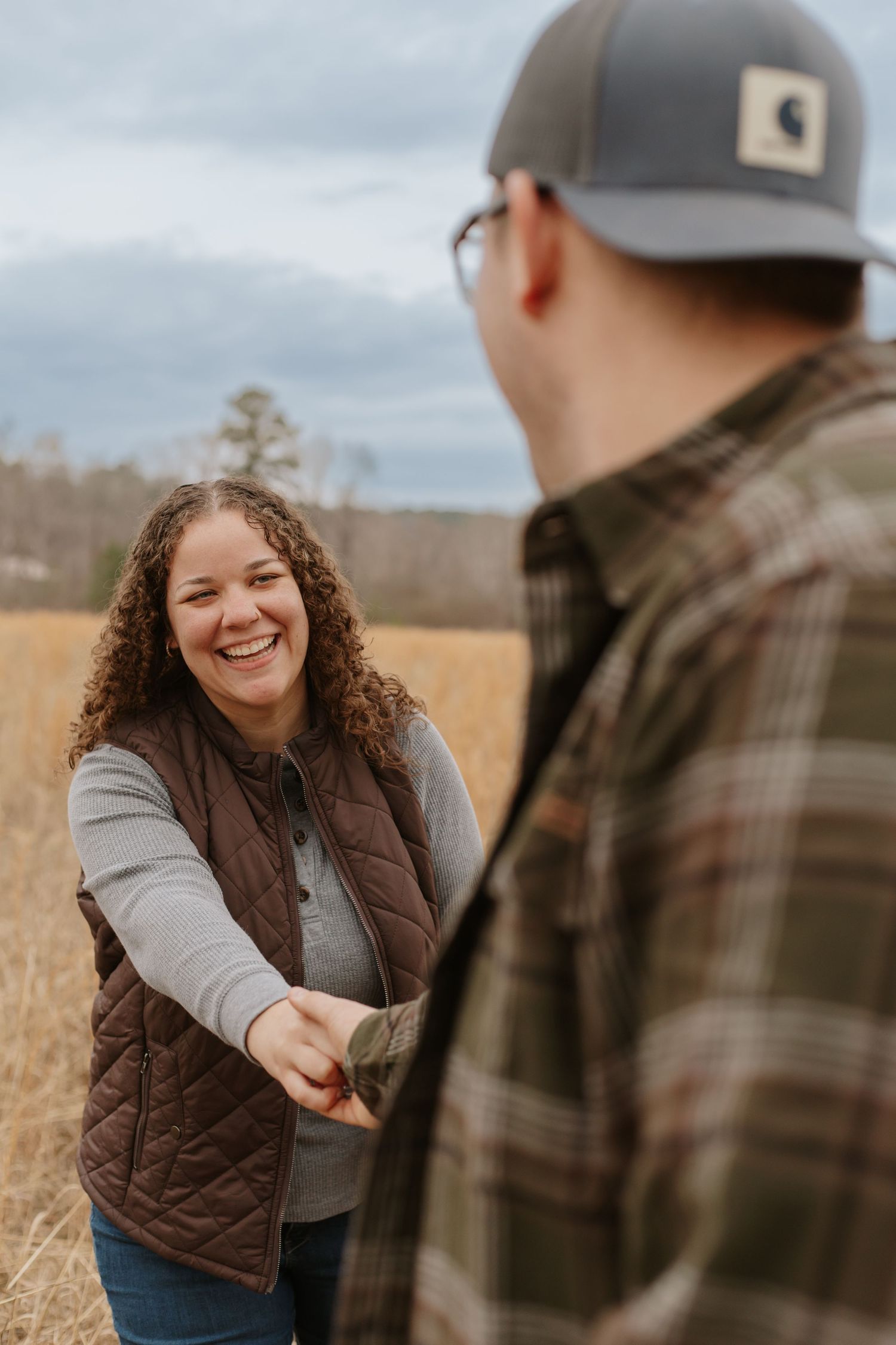Smiling curly-haired woman in brown vest holds hands with man in plaid shirt in autumn field.