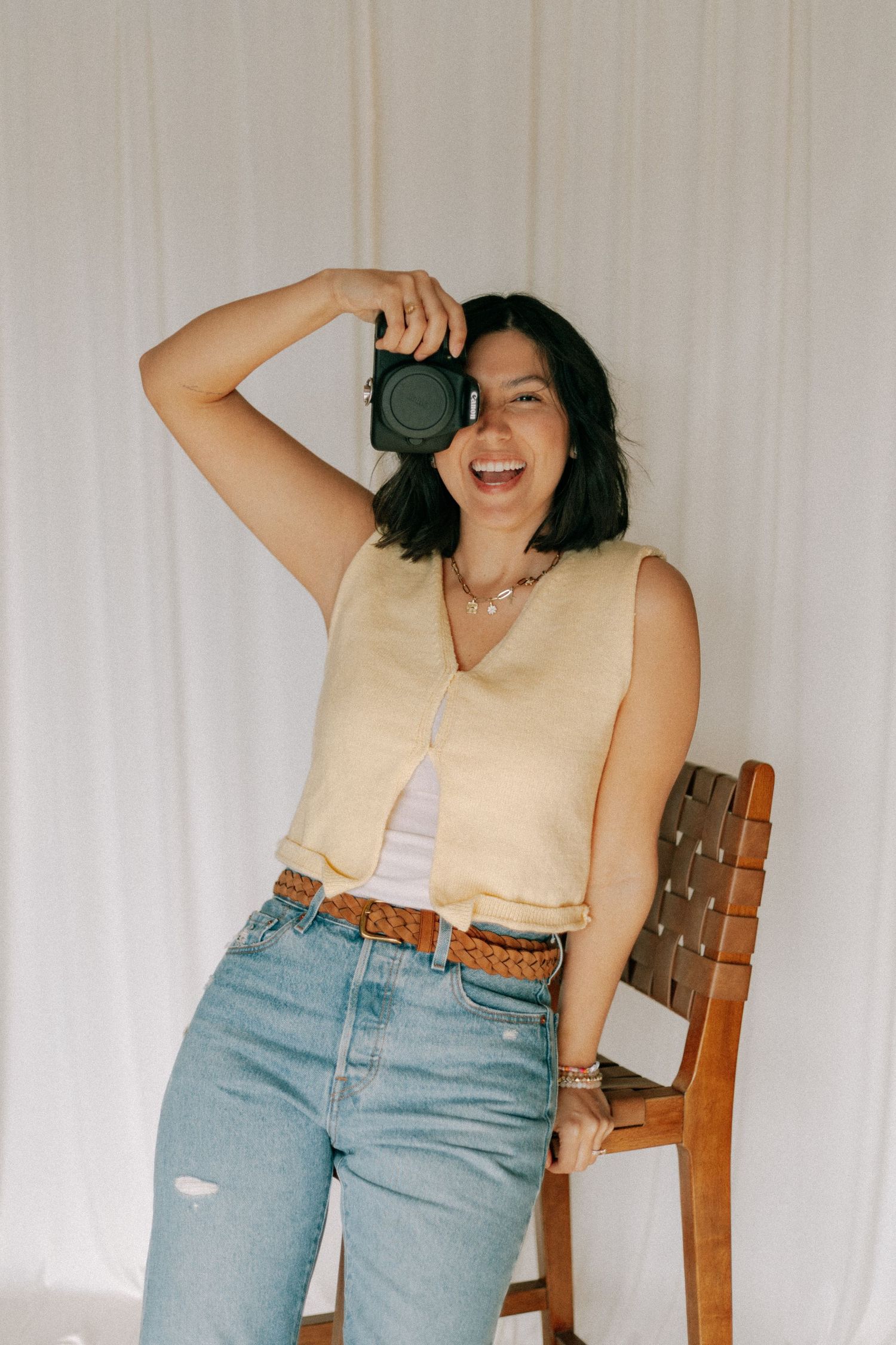 A smiling woman in a yellow vest and jeans holds a camera up to her eye while leaning against a wooden chair.