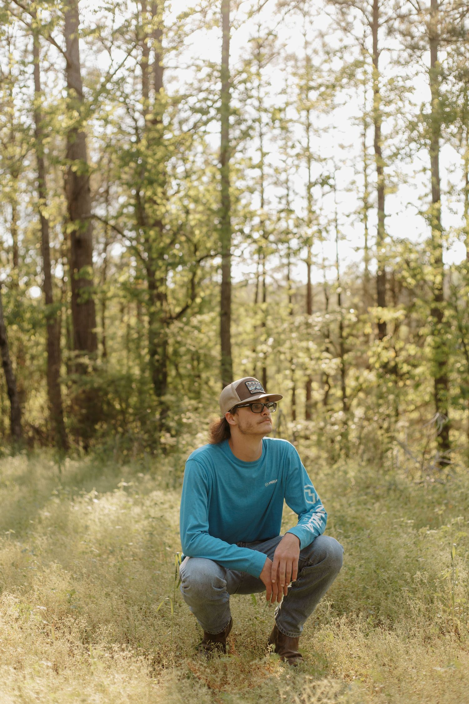 Young man in teal shirt and cap crouches in sunlit forest clearing surrounded by tall trees.