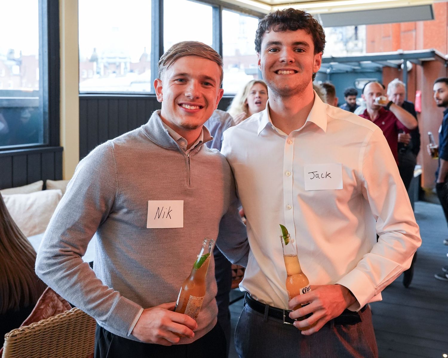 Two young men smiling and holding drinks at a networking event wearing name badges indoors.