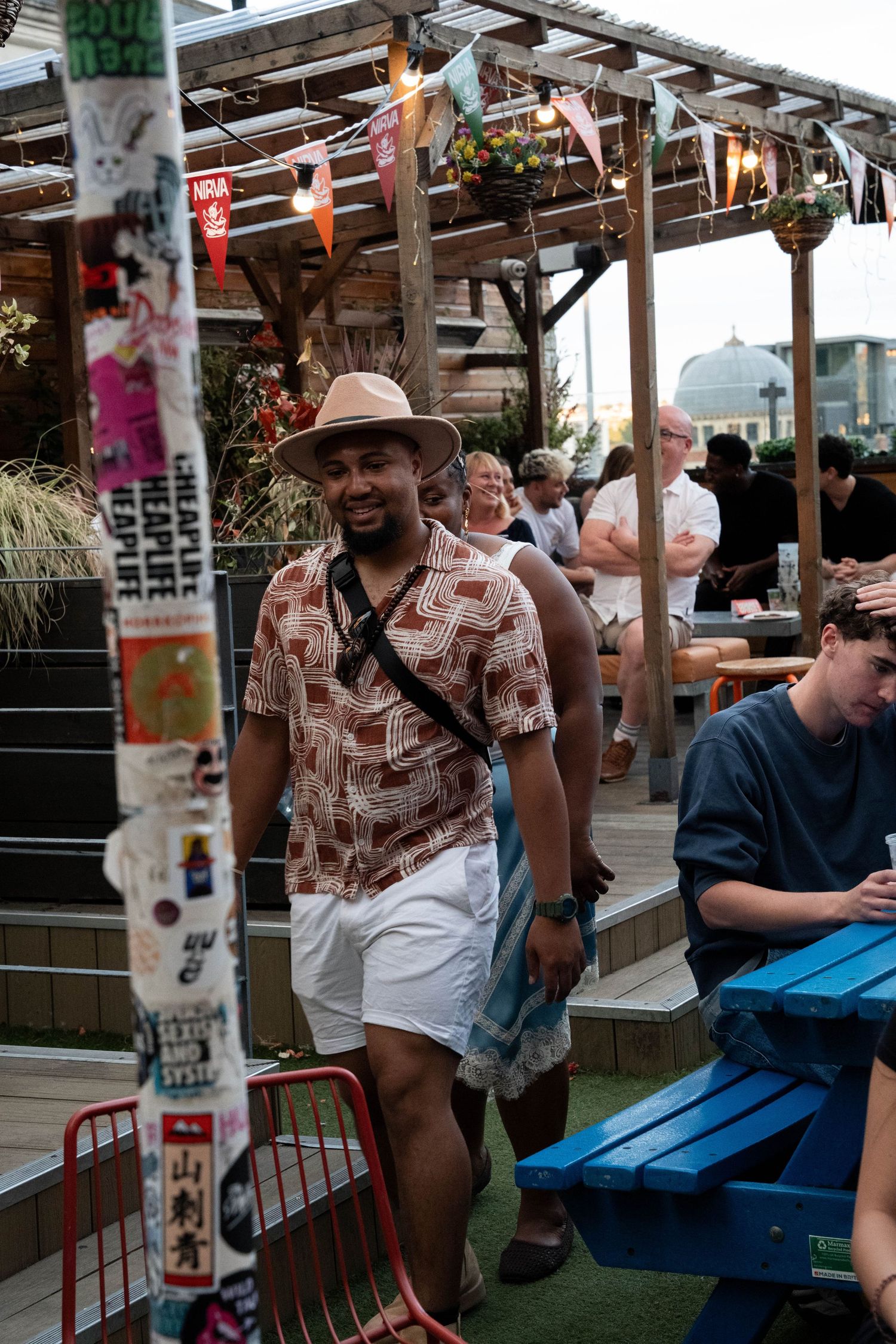 Smiling man in straw hat and patterned shirt at a lively outdoor rooftop bar.