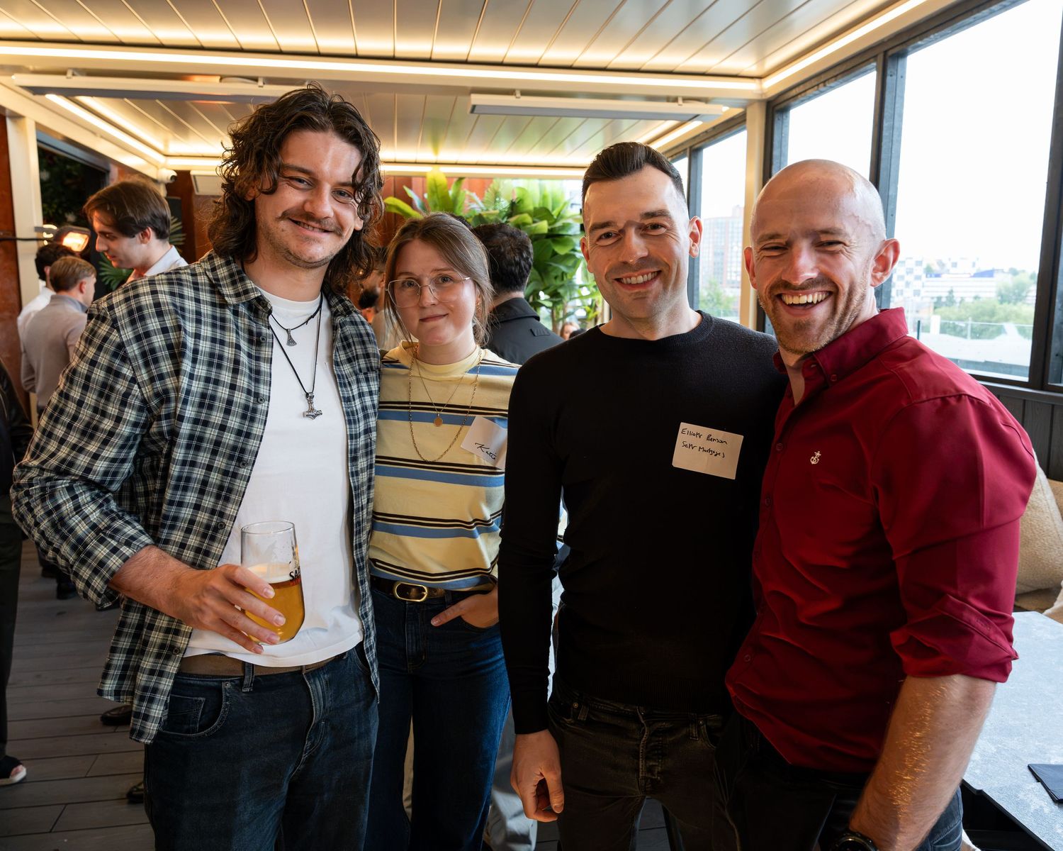 Four men smiling together holding drinks at a casual indoor social networking event with plants nearby.
