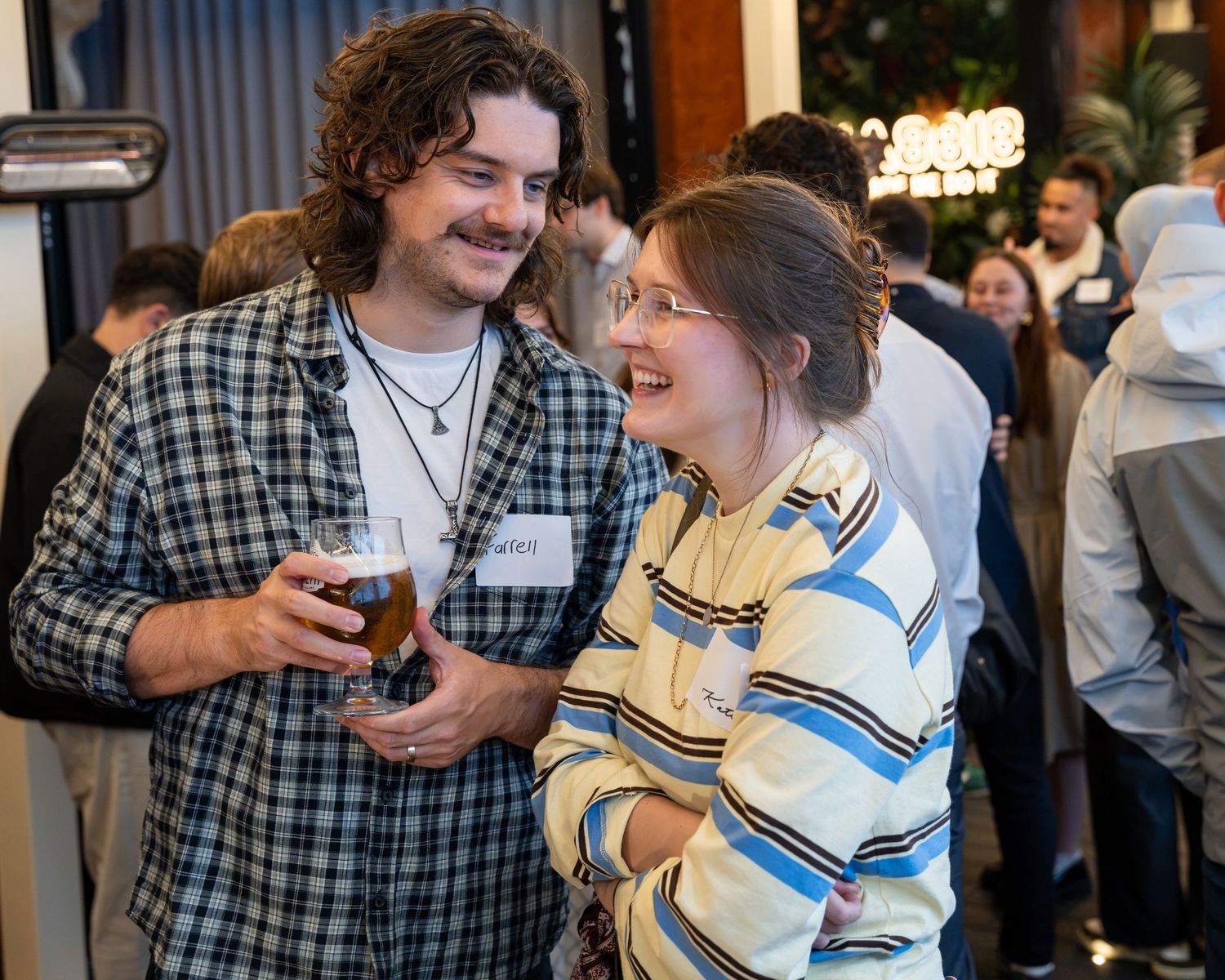 Young man with curly hair holding beer laughing with woman at indoor social event.