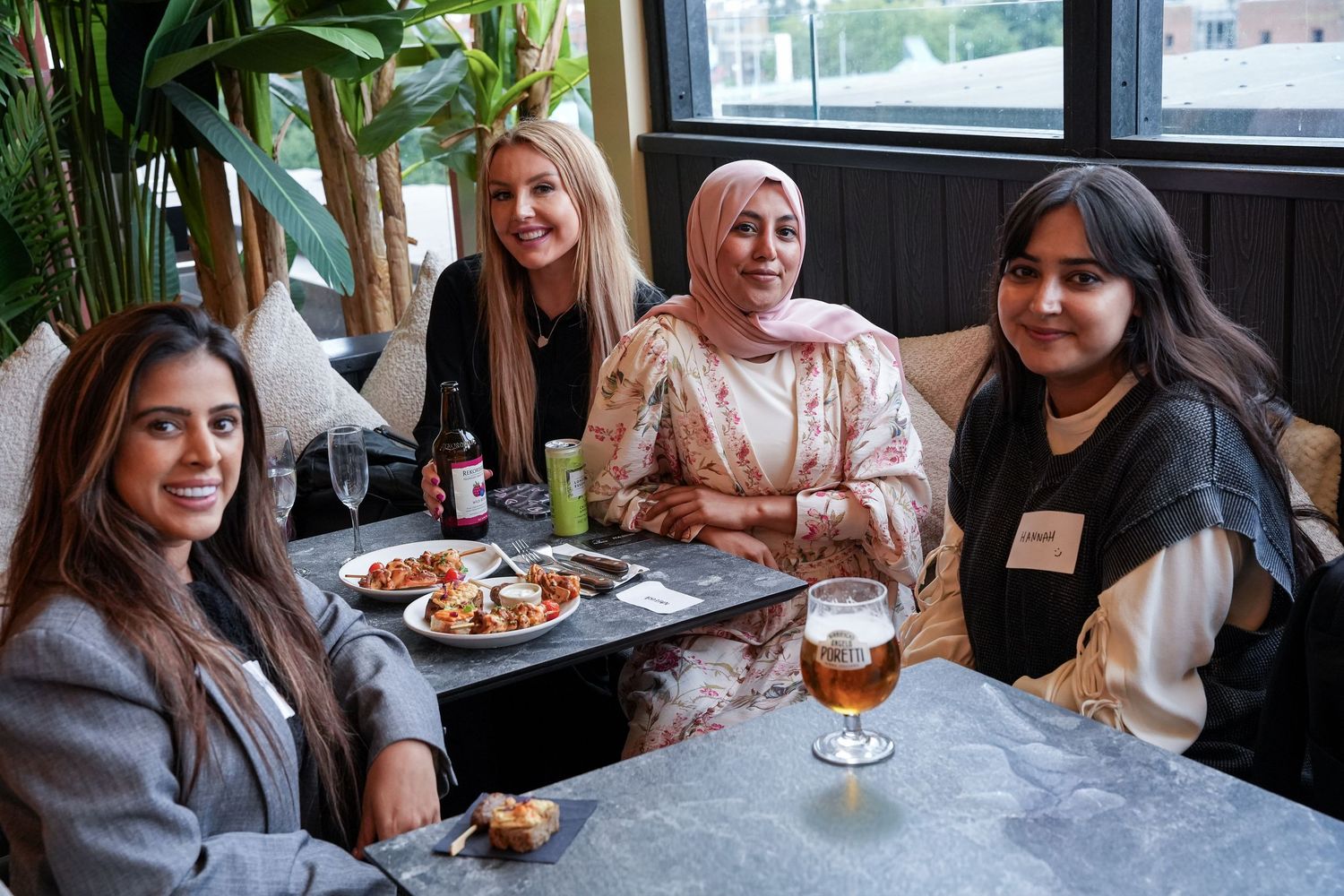 Four women smiling together at a restaurant table with food, drinks, and a beer on display.