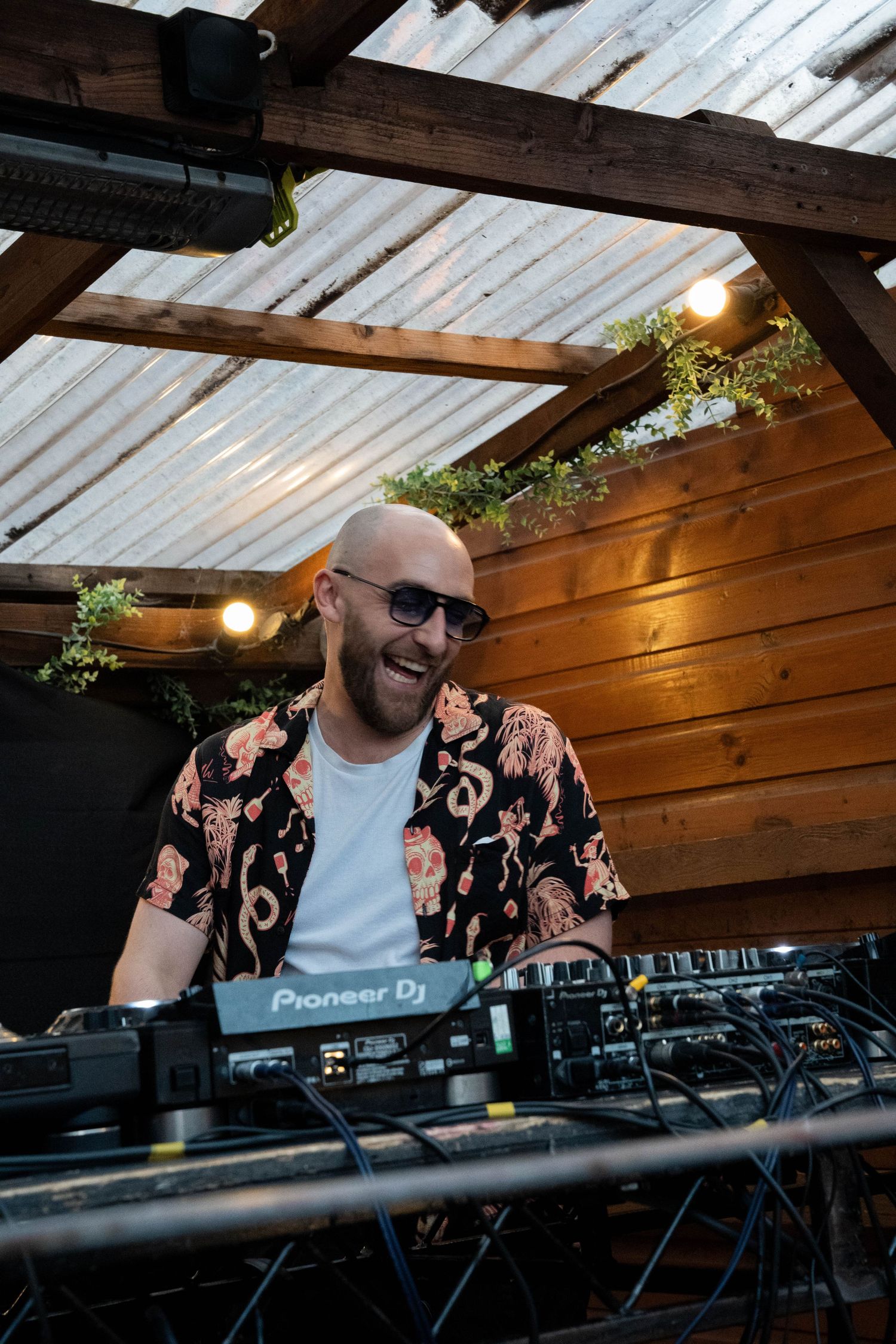 Bald DJ with sunglasses smiling behind Pioneer DJ decks at an outdoor venue.