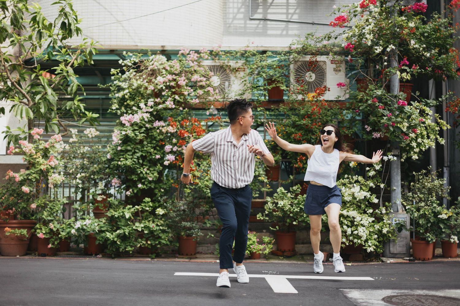 A joyful Asian couple holds hands and runs together on a street surrounded by lush green plants and colorful flowers.