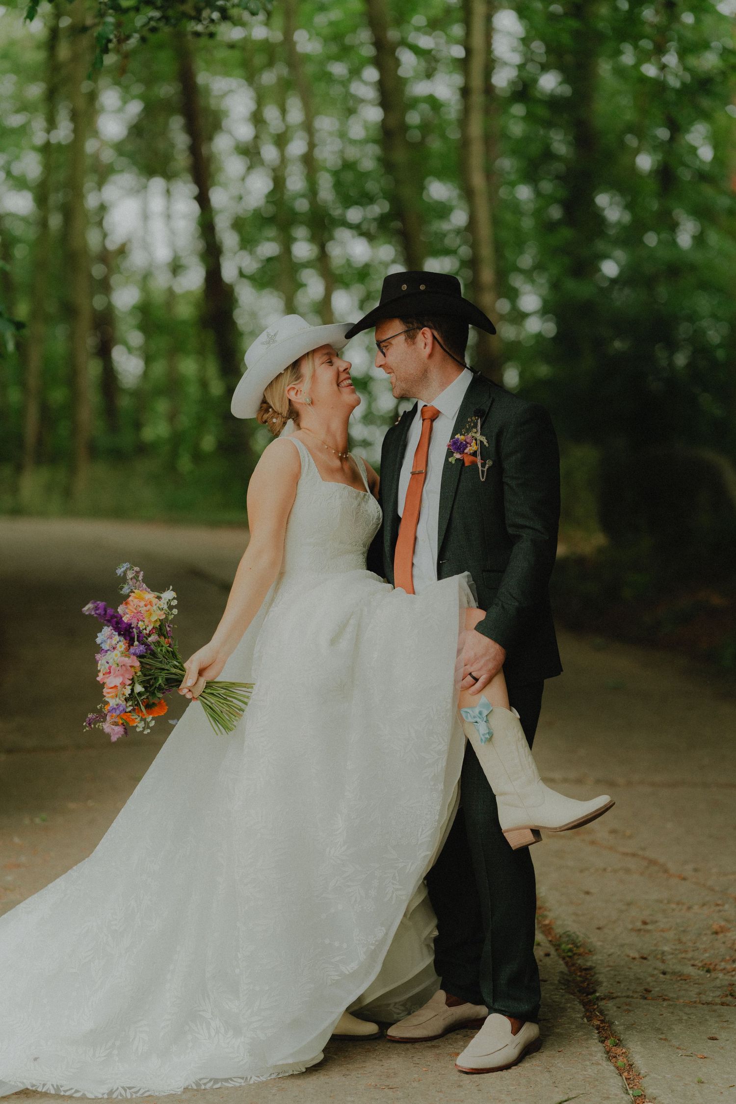 A couple in western-inspired wedding attire embrace on a tree-lined path with a colorful bouquet.