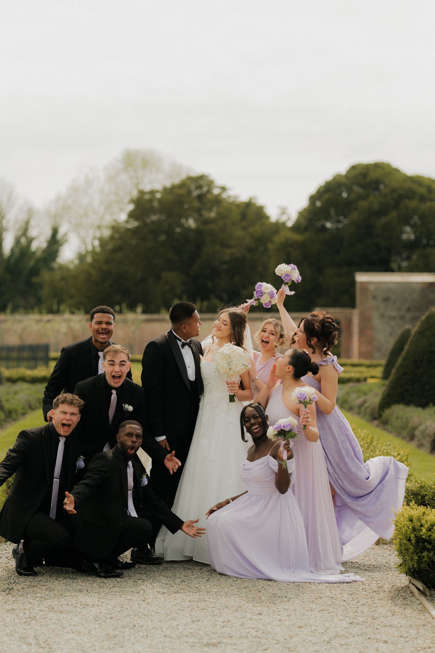 Wedding party celebrating outdoors, bride and groom kissing, bridesmaids in lavender gowns, groomsmen in black suits.