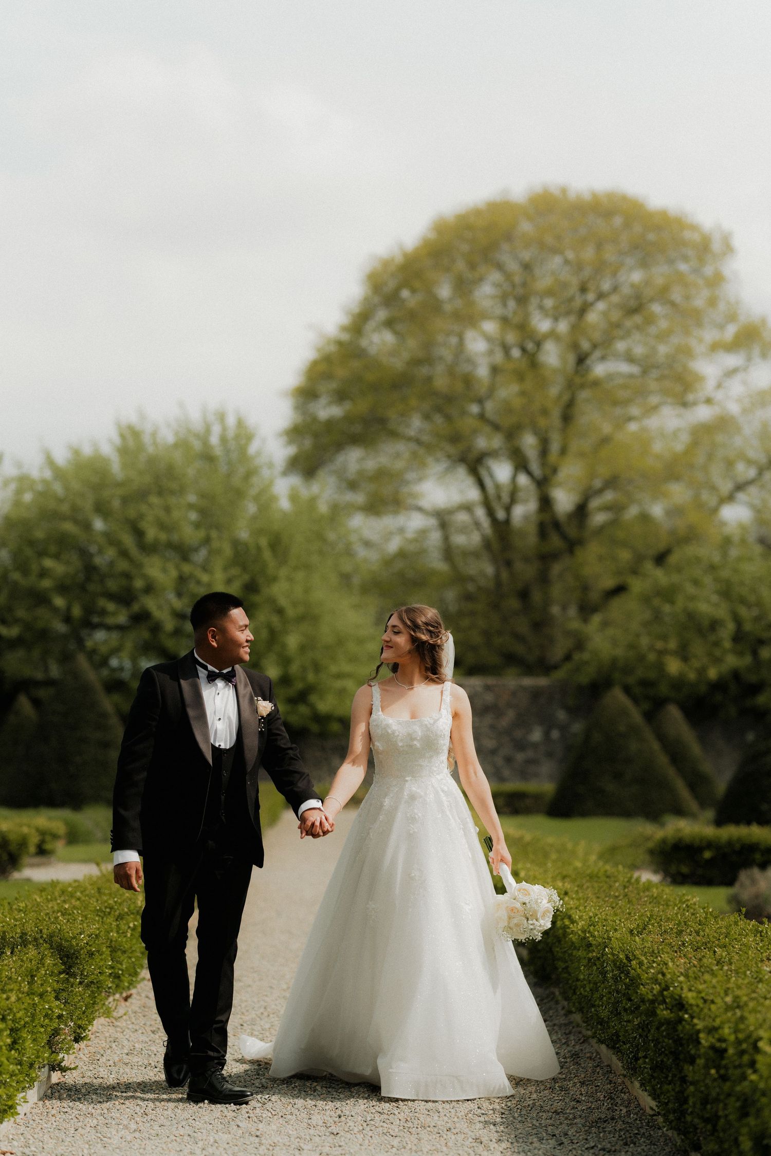 A bride and groom walk hand in hand through a formal garden, smiling at each other on their wedding day.