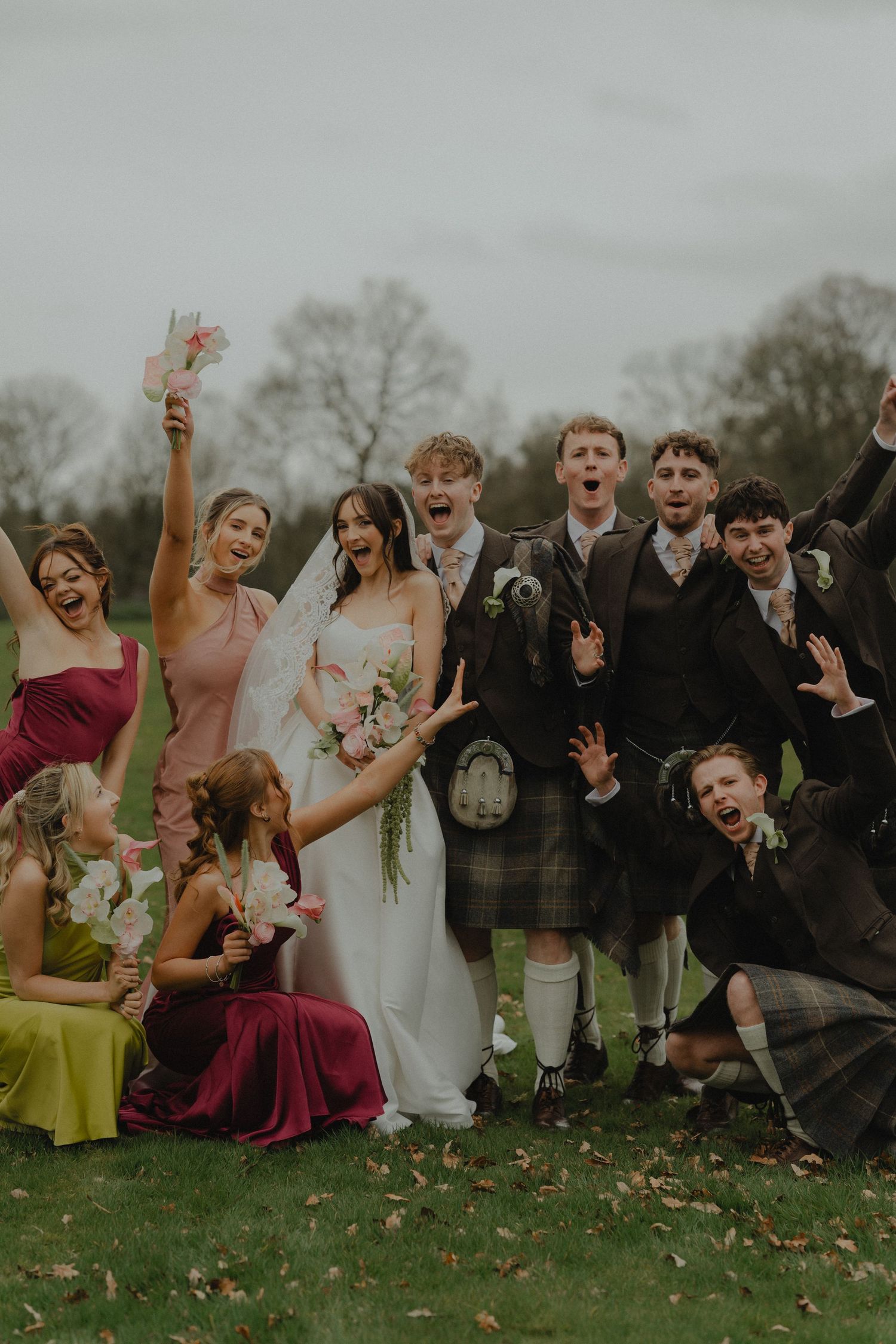 Joyful wedding party celebrating outdoors in colorful bridesmaid dresses and kilts, with bride holding bouquet.