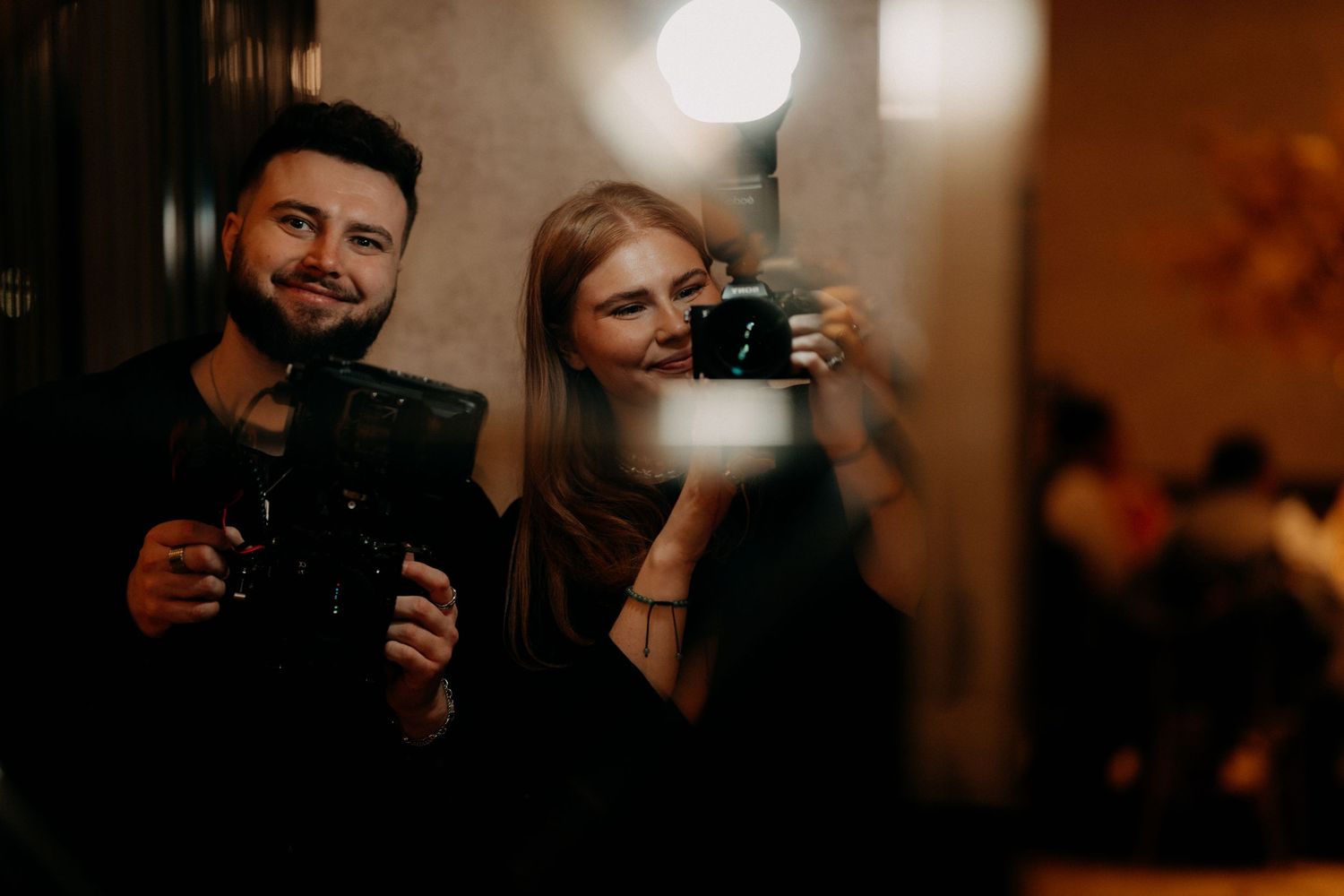 Two photographers holding cameras take a mirror selfie in dimly lit interior.