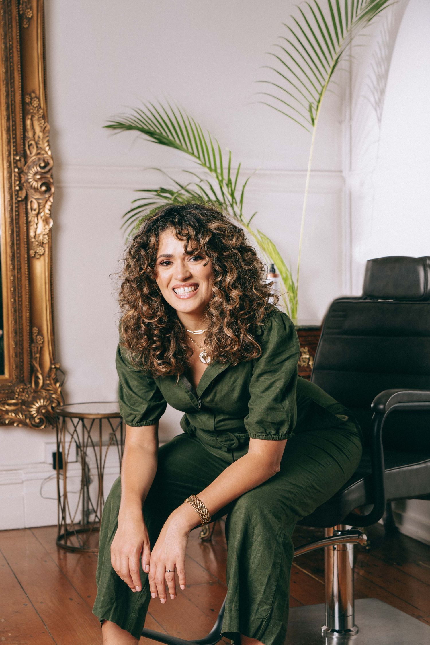 Branding Photography Sessions capture an entrepreneur in a green jumpsuit sitting casually in a bright room with palm fronds and gold frame.