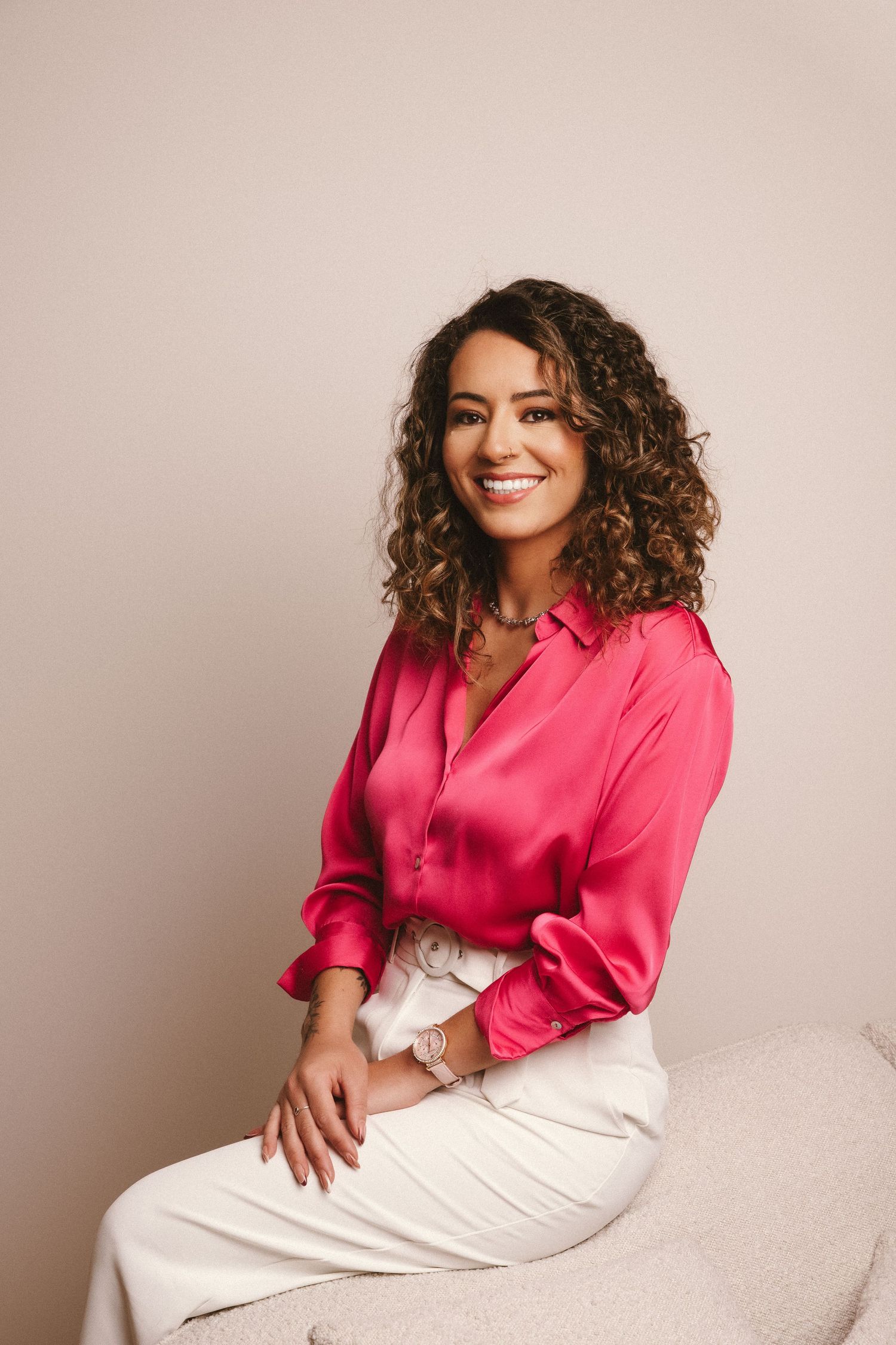 Smiling entrepreneur in a pink silk blouse and white pants sitting casually against a beige background, showcasing professional photo.