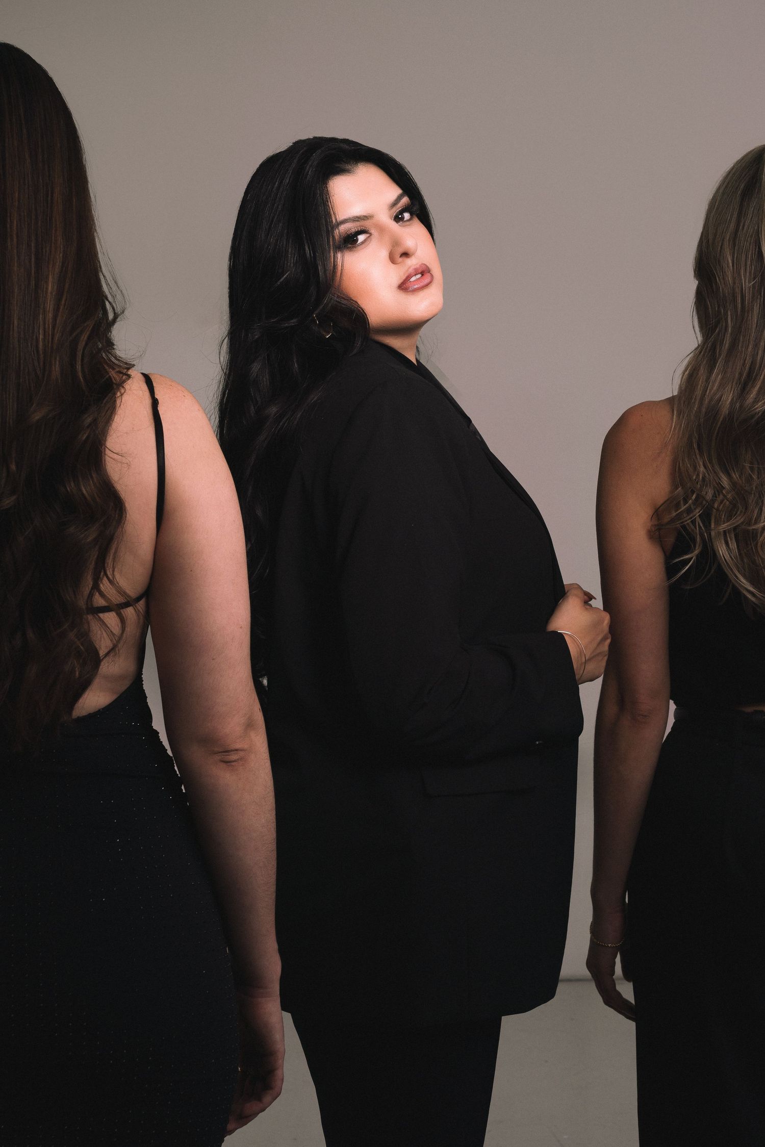 Three women in black outfits pose together, with the center woman in a blazer looking over her shoulder.