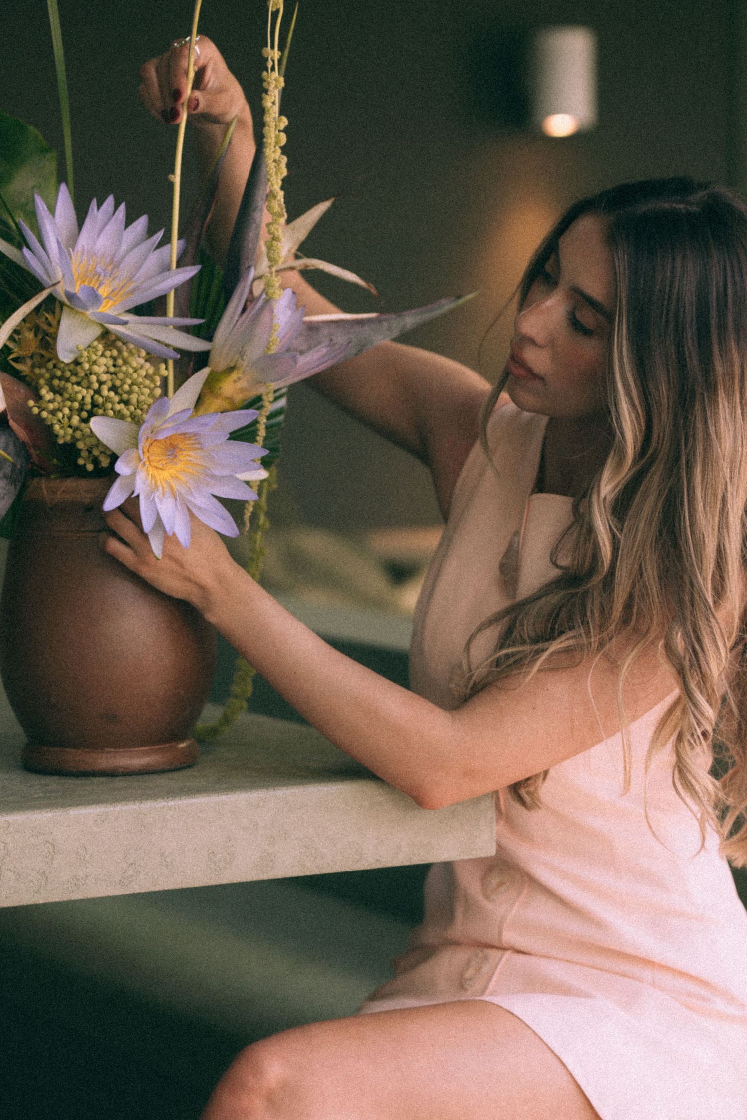 Personal branding photoshoot in Sydney captures an entrepreneur in a pink dress arranging purple water lilies and wheat stalks in dim light.