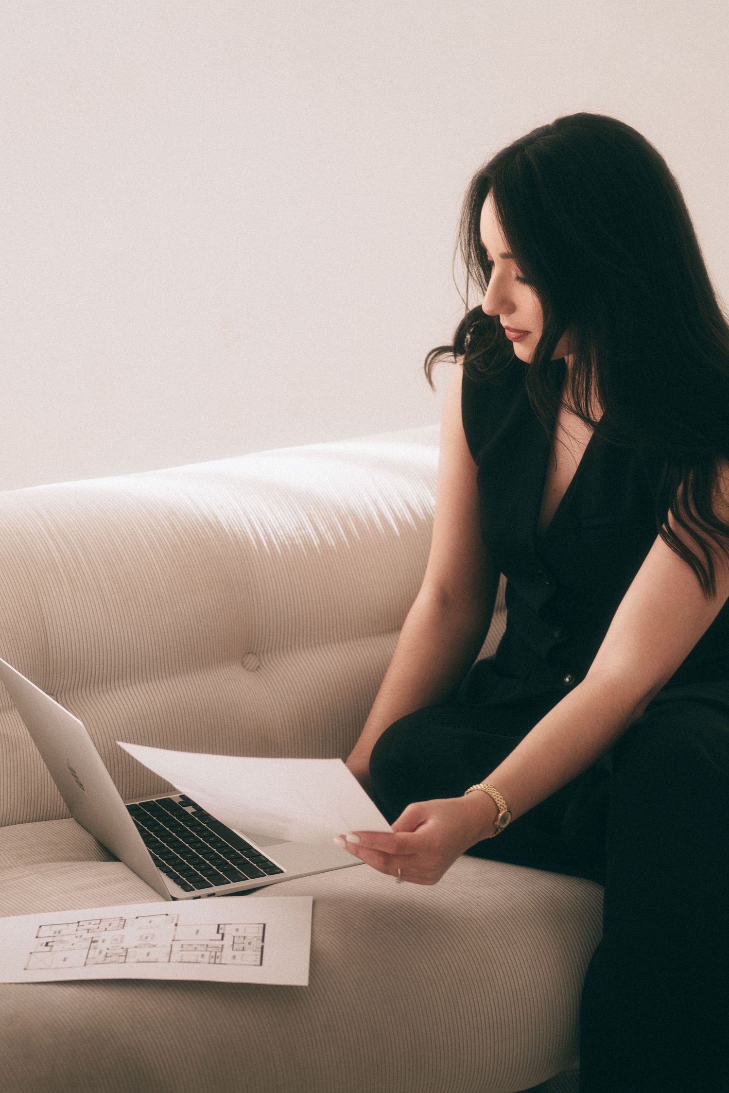 Branding Photography Sessions capture an entrepreneur in a black dress working on a laptop with papers on a beige couch in a minimalist.