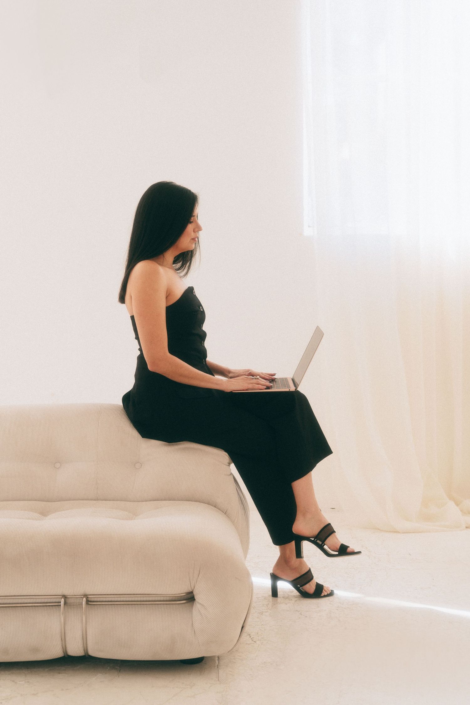 Sophisticated coach capture an entrepreneur in a black dress working on a laptop while sitting on a beige couch in a minimal white room.