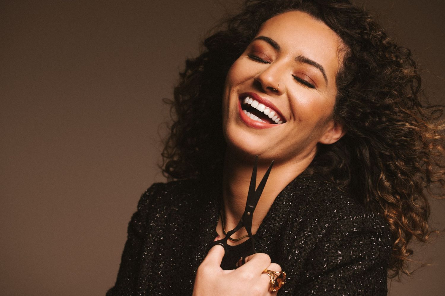 Joyful entrepreneur with curly hair wearing a black sparkly top laughs with eyes closed against a brown background.