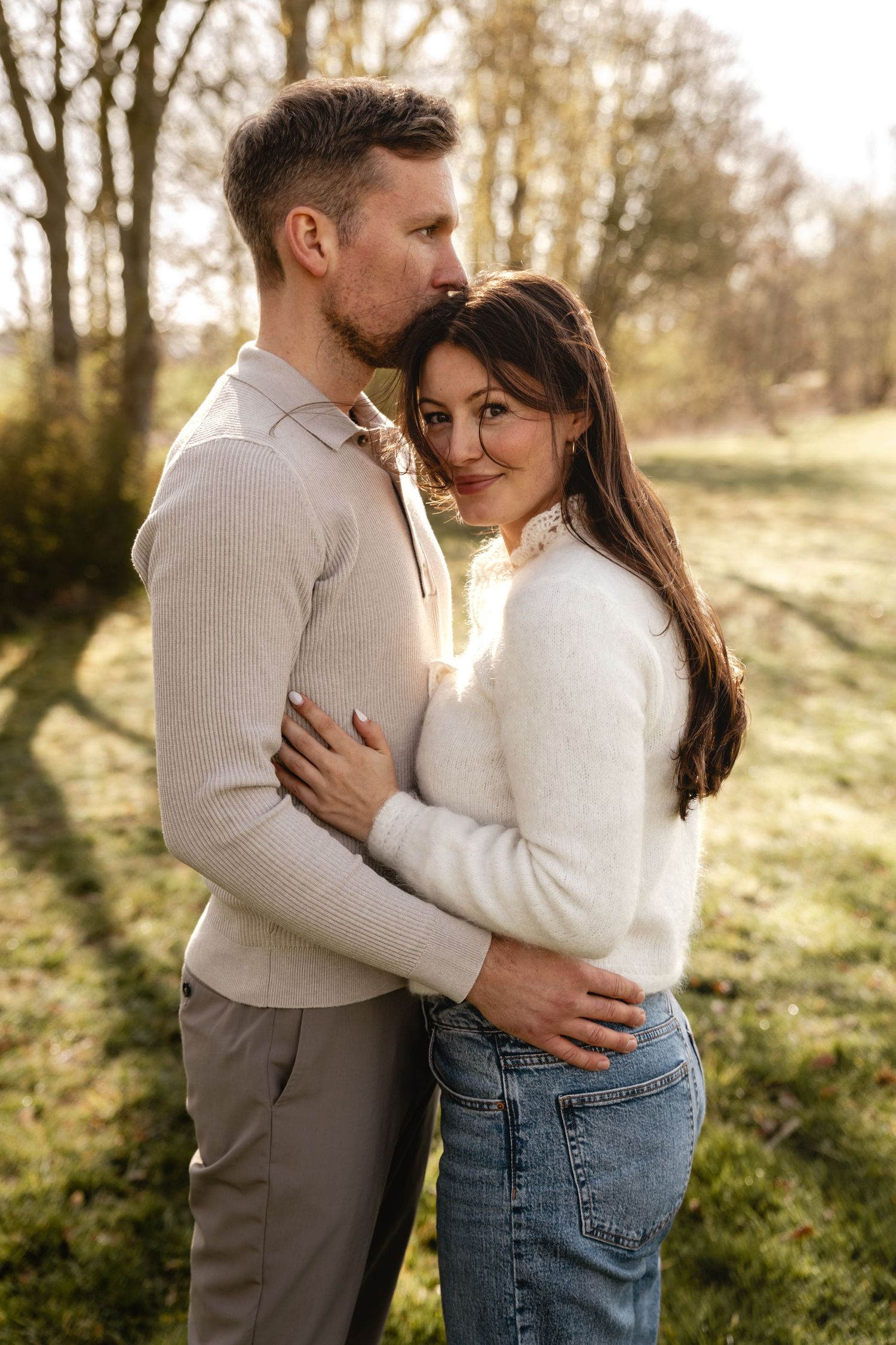 Lächelnde Frau im weißen Strickpullover wird von ihrem Partner auf einer sonnigen Wiese umarmt und auf die Stirn geküsst.