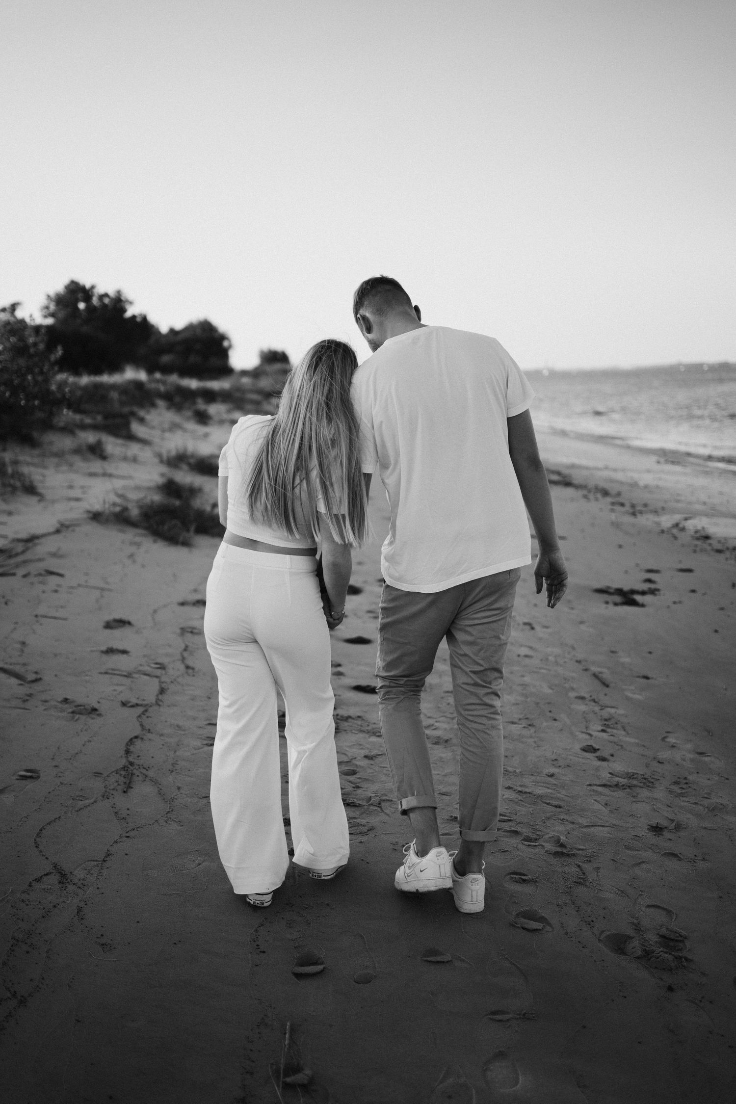 Black and white photo of a couple in casual white clothes walking arm in arm along a sandy beach shoreline.