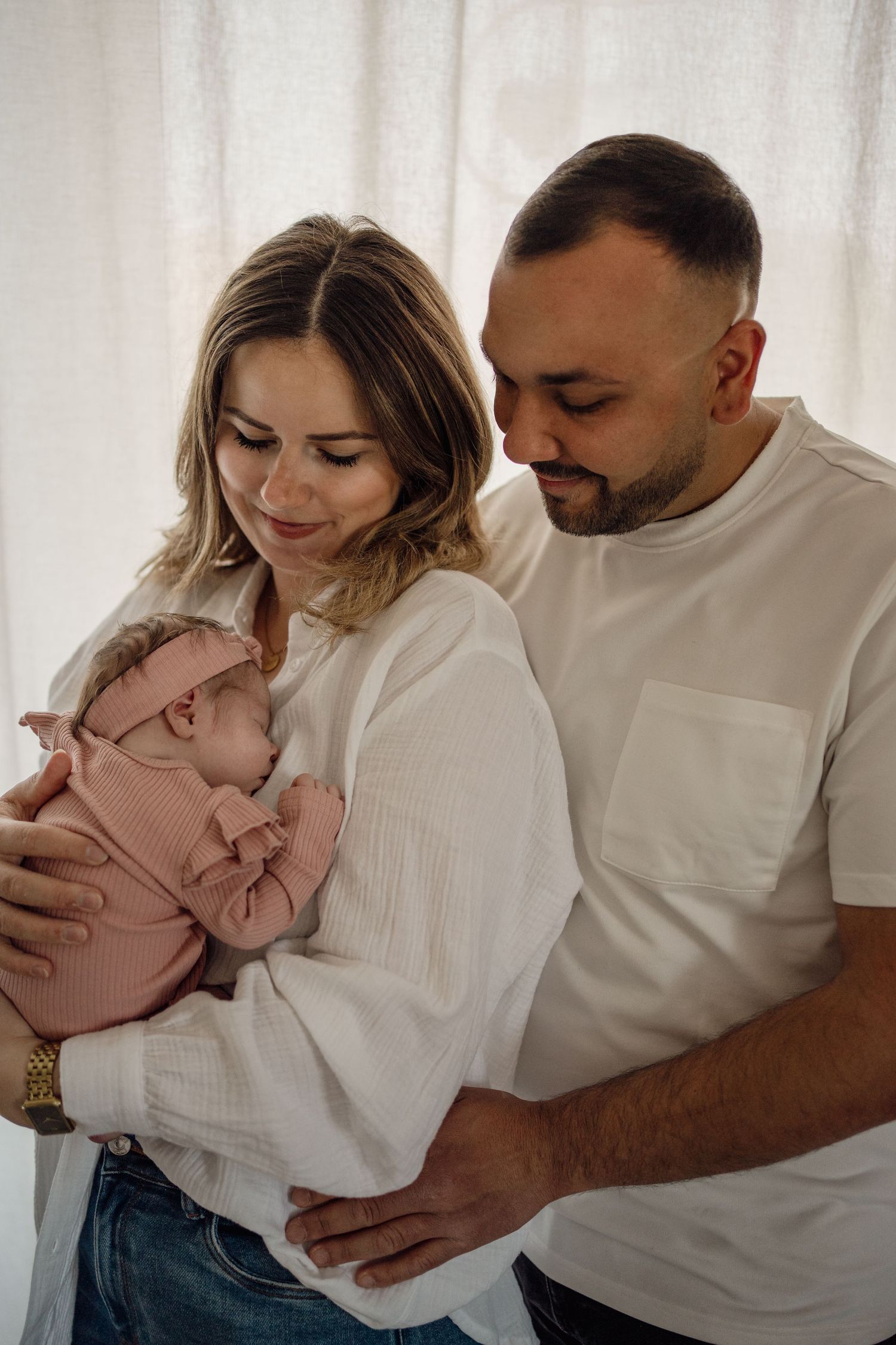 A smiling mother in a white blouse cradles her sleeping newborn in pink while the father embraces them both.
