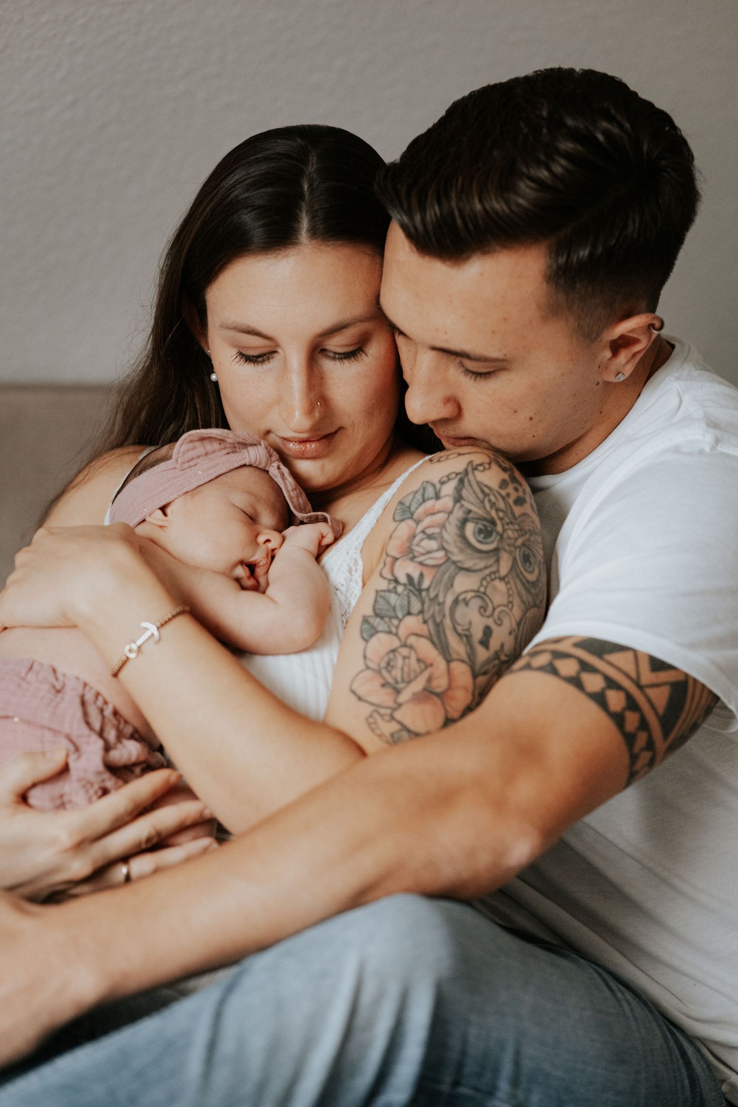 Tätowierte Mutter und Vater halten ihr schlafendes Neugeborenes mit rosa Haarband während eines Familien-Fotoshootings liebevoll im Arm.