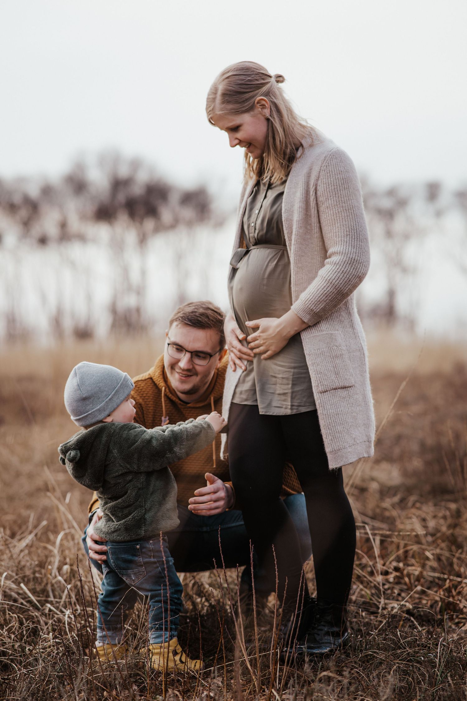 Expecting mother in a cozy cardigan with her husband and toddler in a golden autumn field during a maternity photo session.